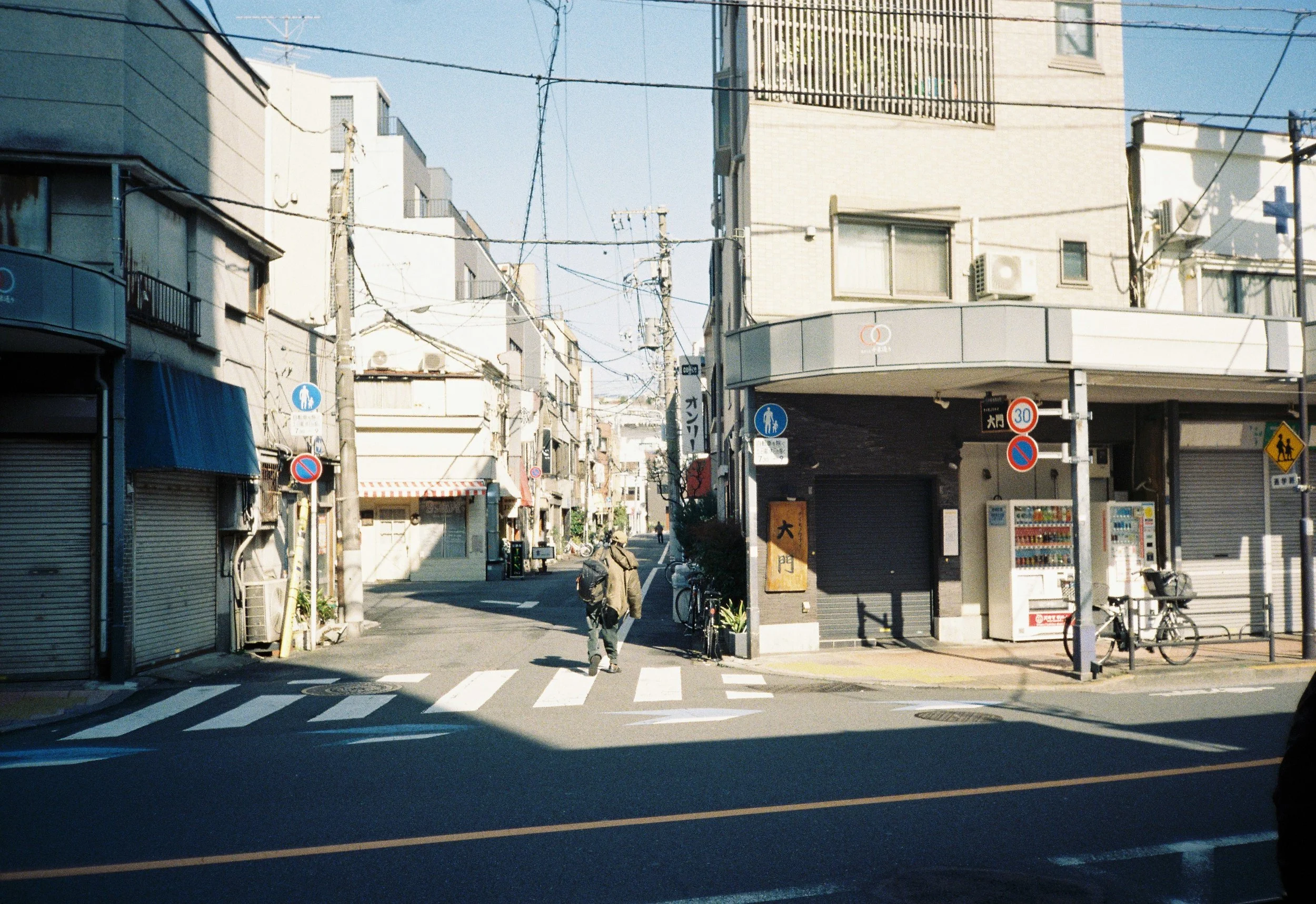 Vista de una calle urbana en Japón con edificios de varios pisos, cables eléctricos y señales de tránsito. Hay una persona cruzando la calle con una bicicleta en un cruce peatonal.