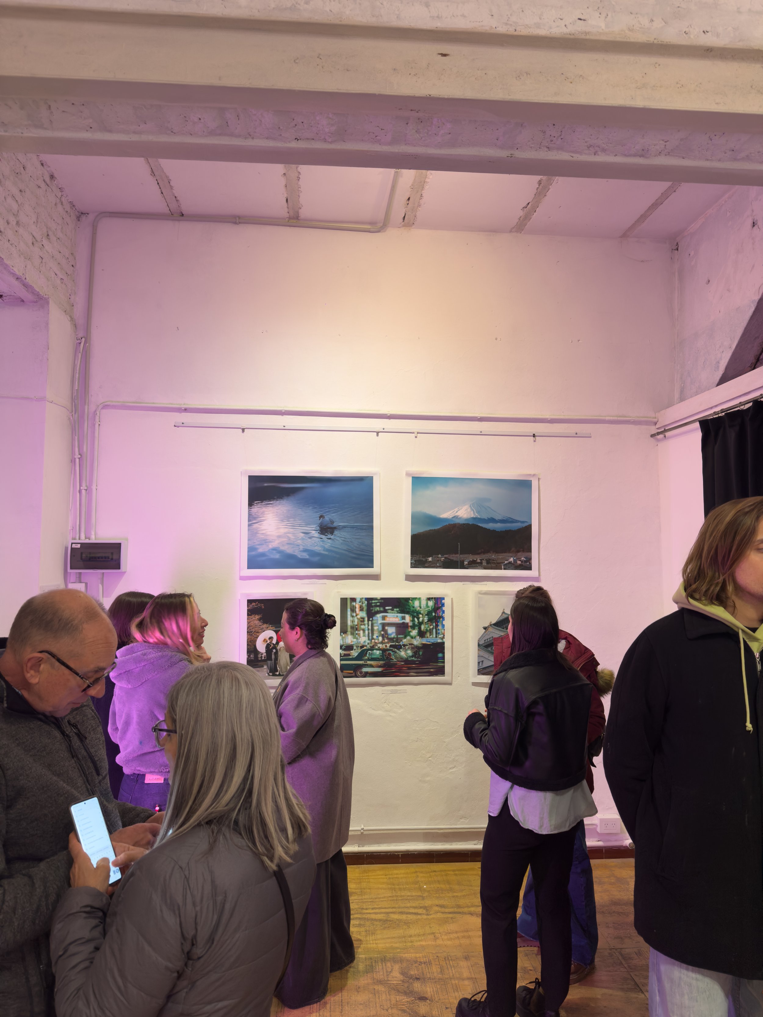 Vista de una galería de arte con varias personas observando fotografías en la pared, fondo colorido con iluminación suave.