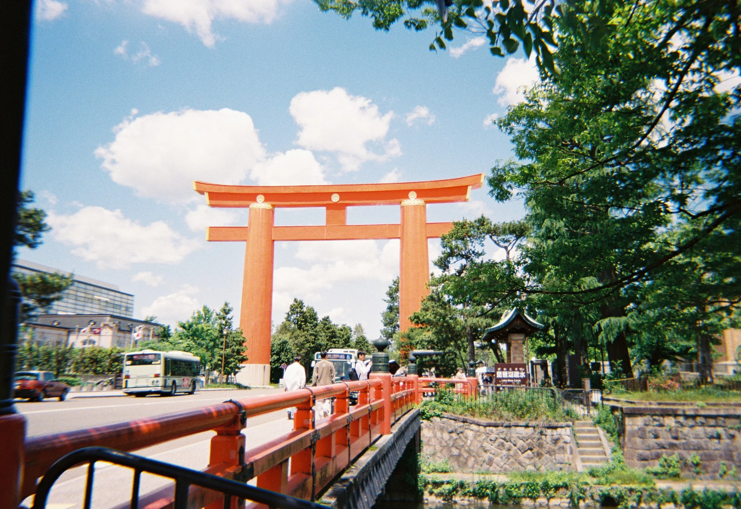 Un torii rojo tradicional japonés en un entorno urbano con árboles y edificios alrededor, y un cielo azul con nubes.