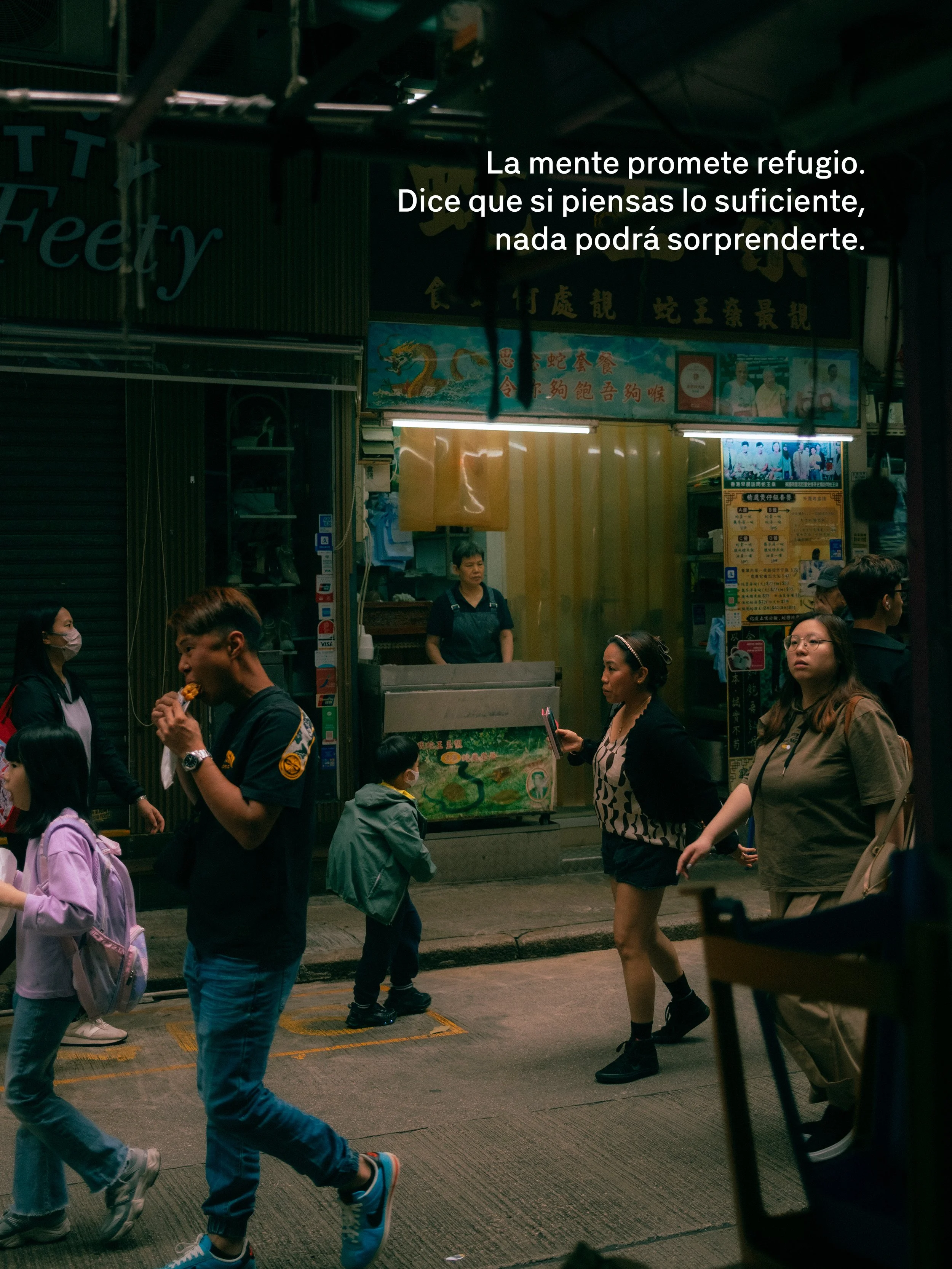 Personas caminando en una calle concurrida frente a un puesto de comida o tienda en un área urbana, con carteles y señalización en chino y luces fluorescentes.