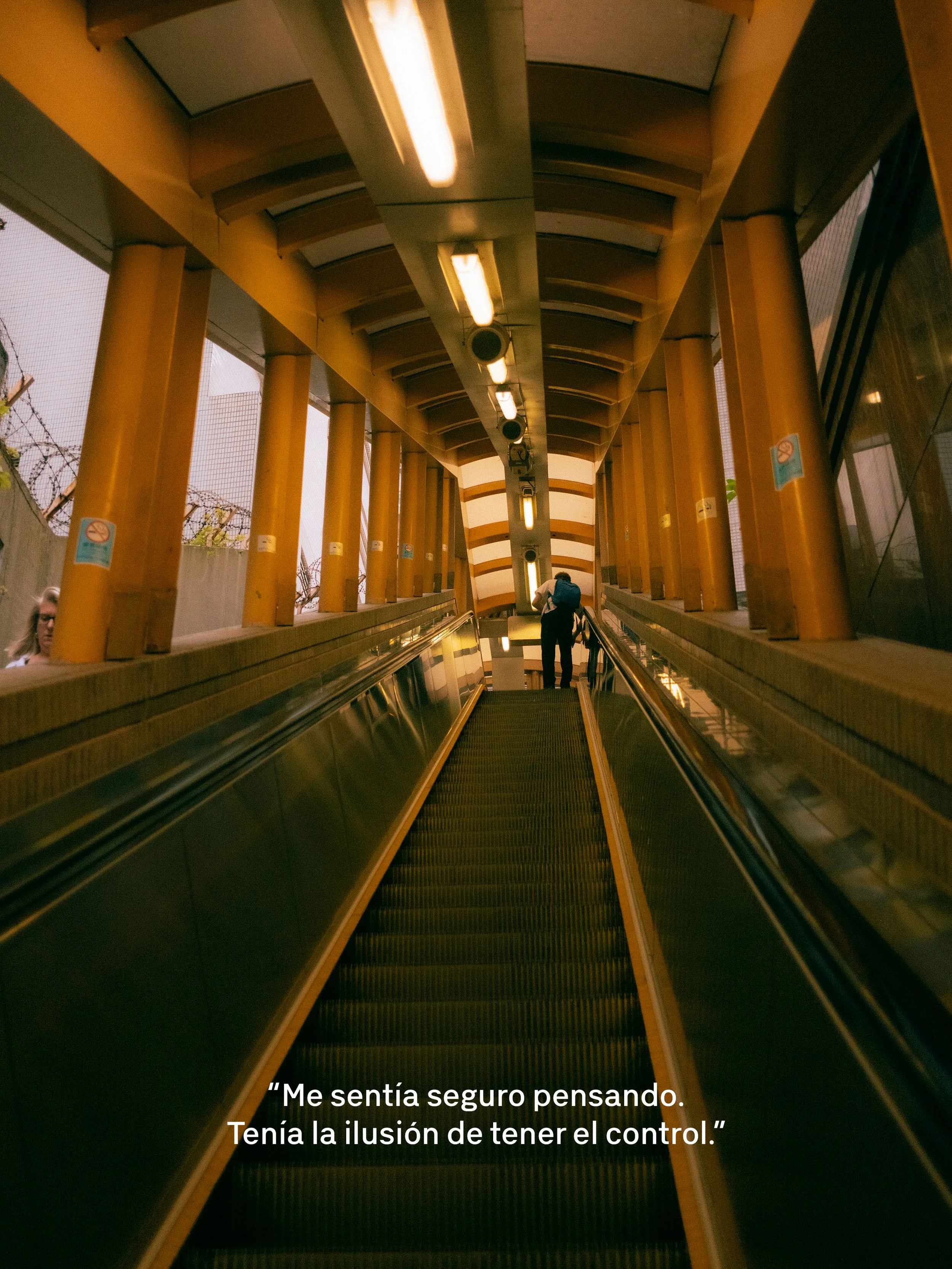 Perspectiva desde abajo de una escalera mecánica en un pasillo con paredes amarillas y ventanas con malla de seguridad. Una persona está subiendo con una mochila.