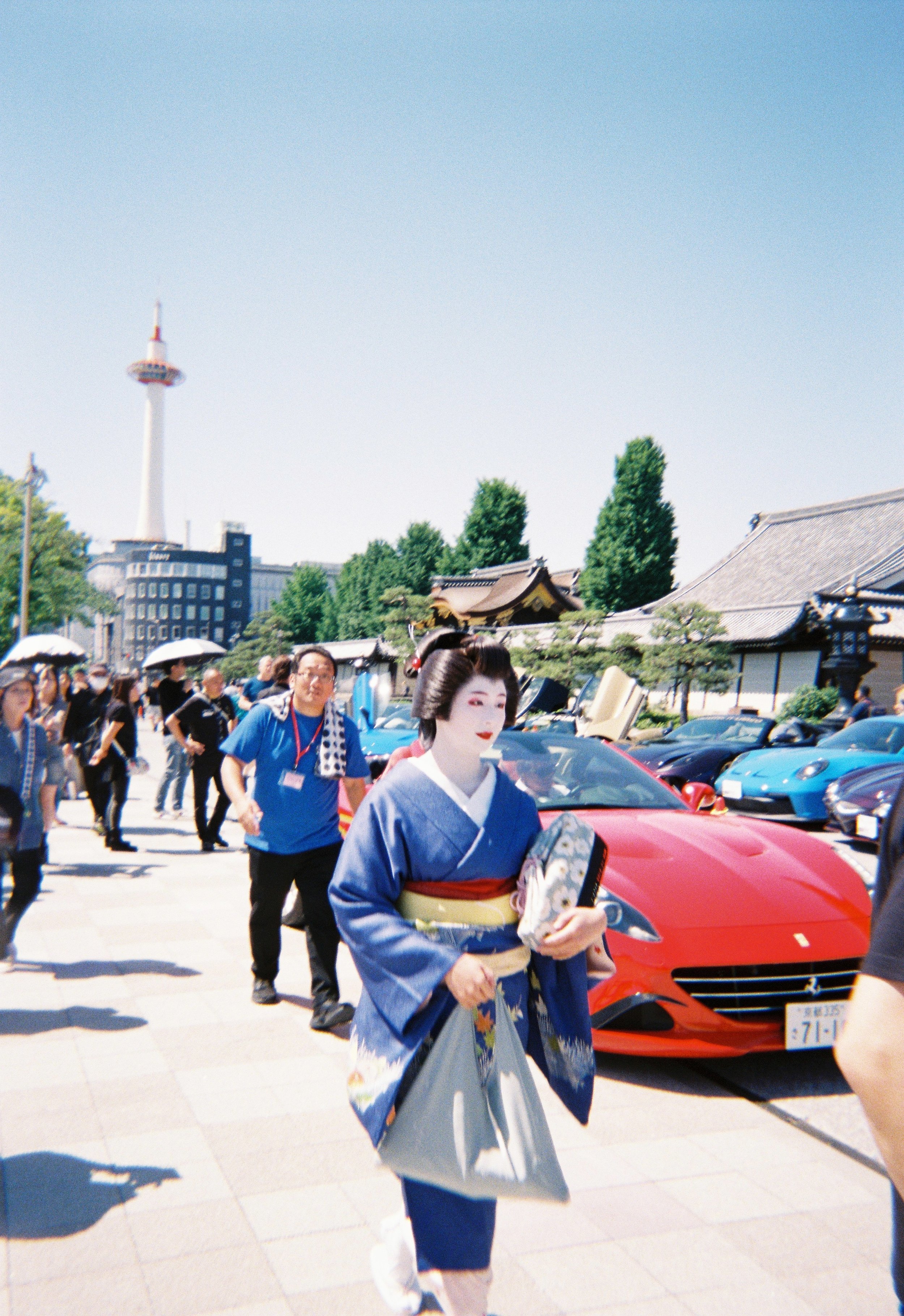 Mujer vestida con kimono paseando entre coches deportivos en una plaza con árboles y edificios, en un día soleado y despejado.