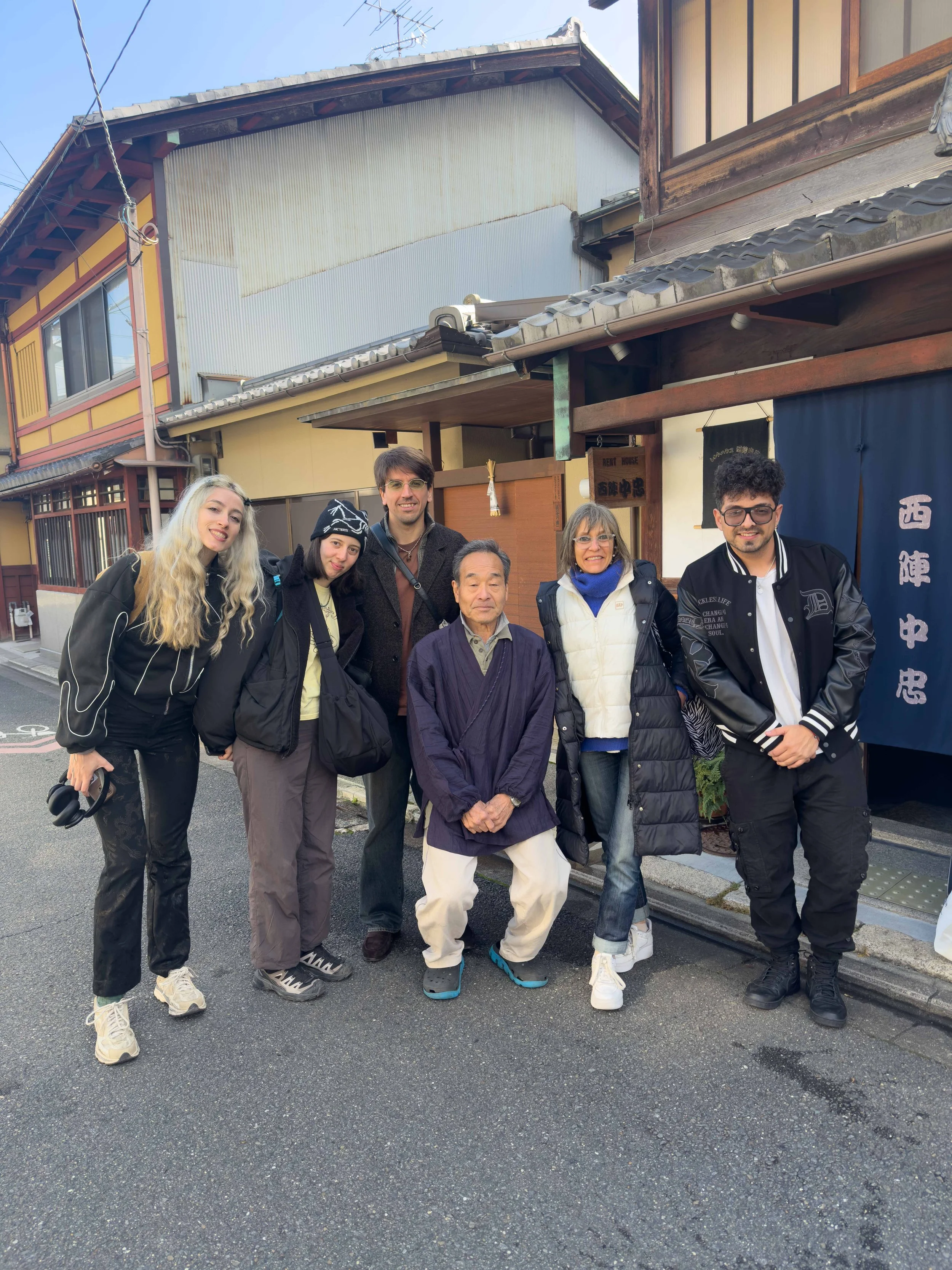 Grupo de seis personas posando en la calle frente a edificios tradicionales de Japón, con paredes de madera, ventanas y puertas de tela, en un día despejado.