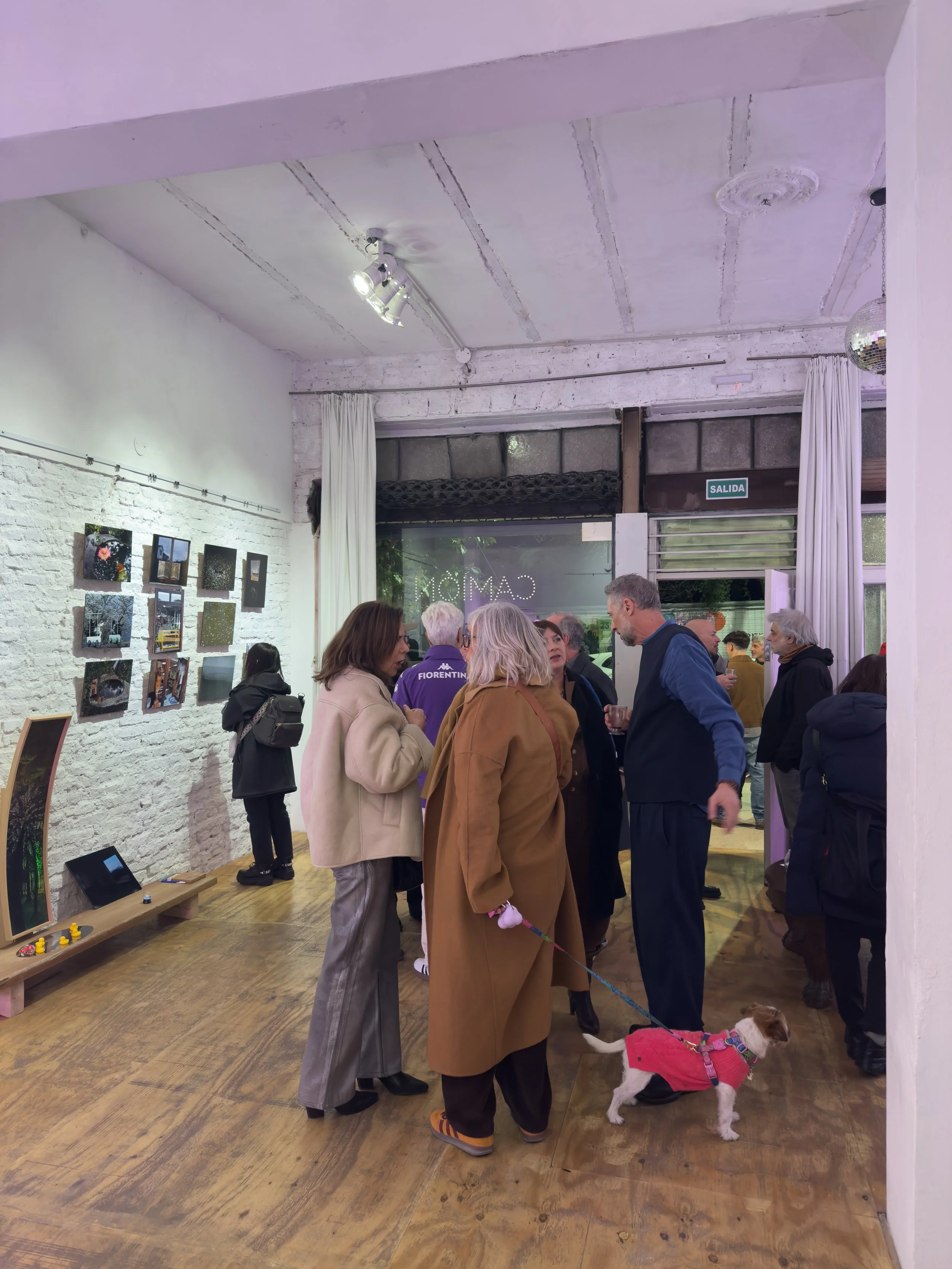 Grupo de personas en un interior de galería de arte, conversando y con un perro pequeño con ropa roja.