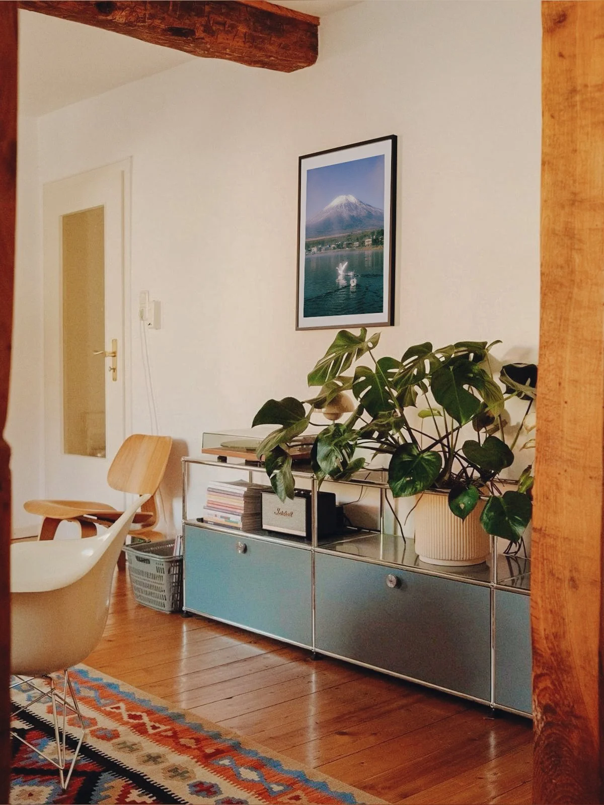 Interior de una sala de estar con un mueble azul, una planta en una maceta, una mesa con libros y una pintura de un volcán en la pared.