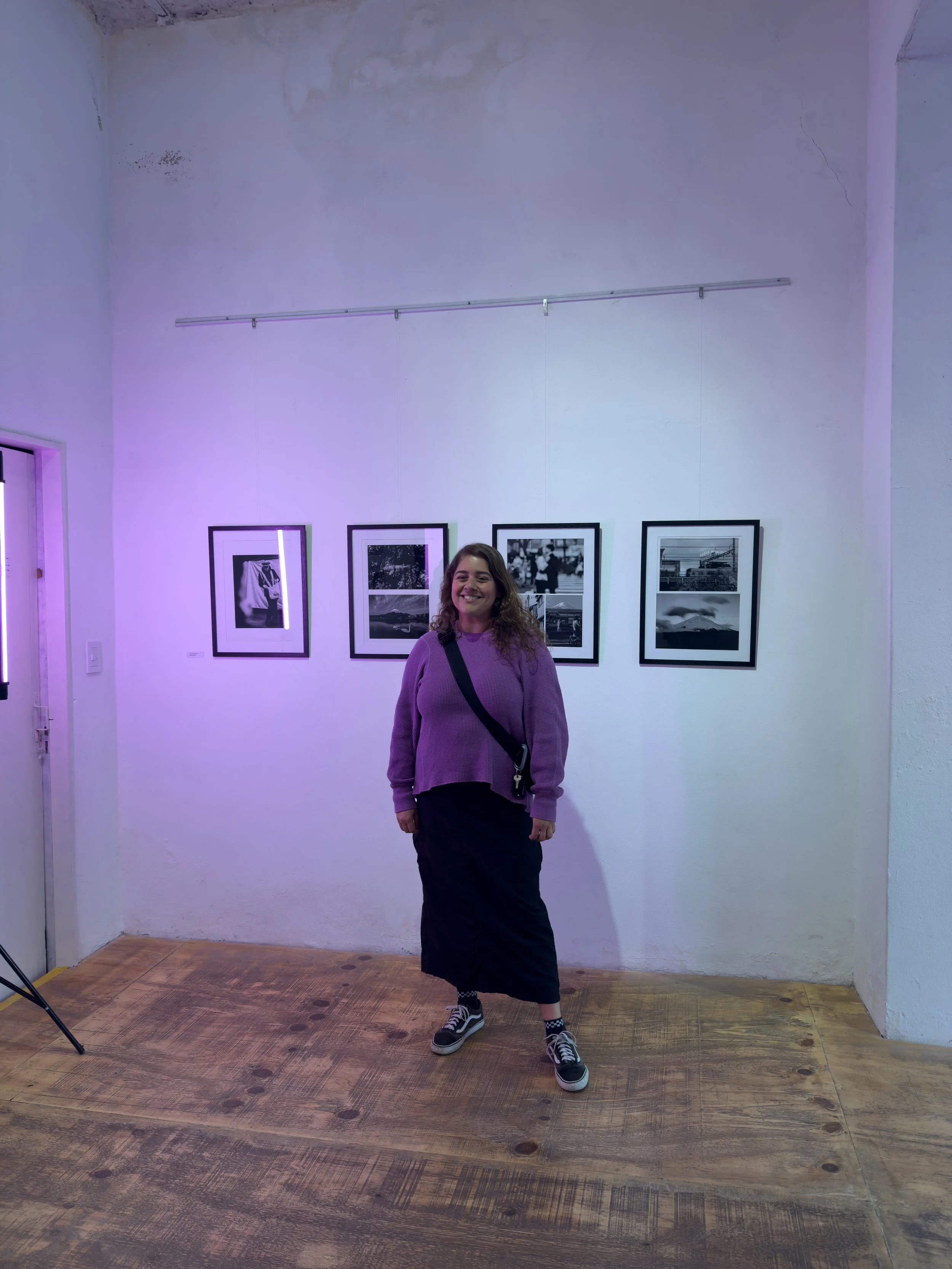 Mujer sonriendo, vistiendo suéter lila, falda larga negra, zapatillas deportivas con calcetas a cuadros, en una galería de arte con fotografías enmarcadas en la pared y piso de madera.
