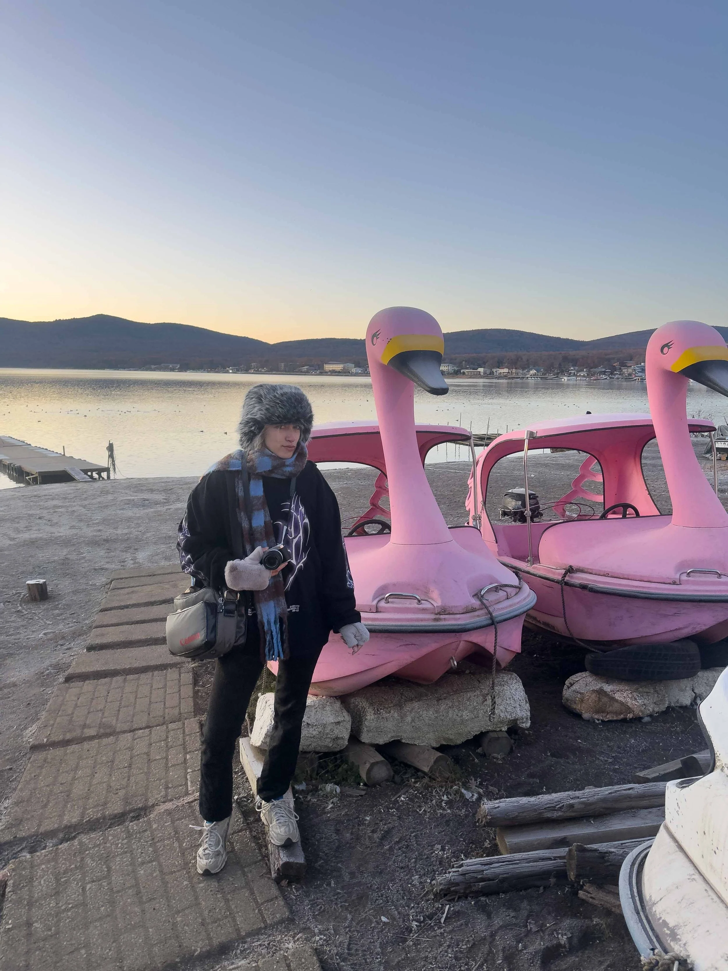 Persona con abrigo y gorro de piel de frente a barcos en forma de cisne en un muelle, con vista a un lago con montañas al fondo durante el atardecer.