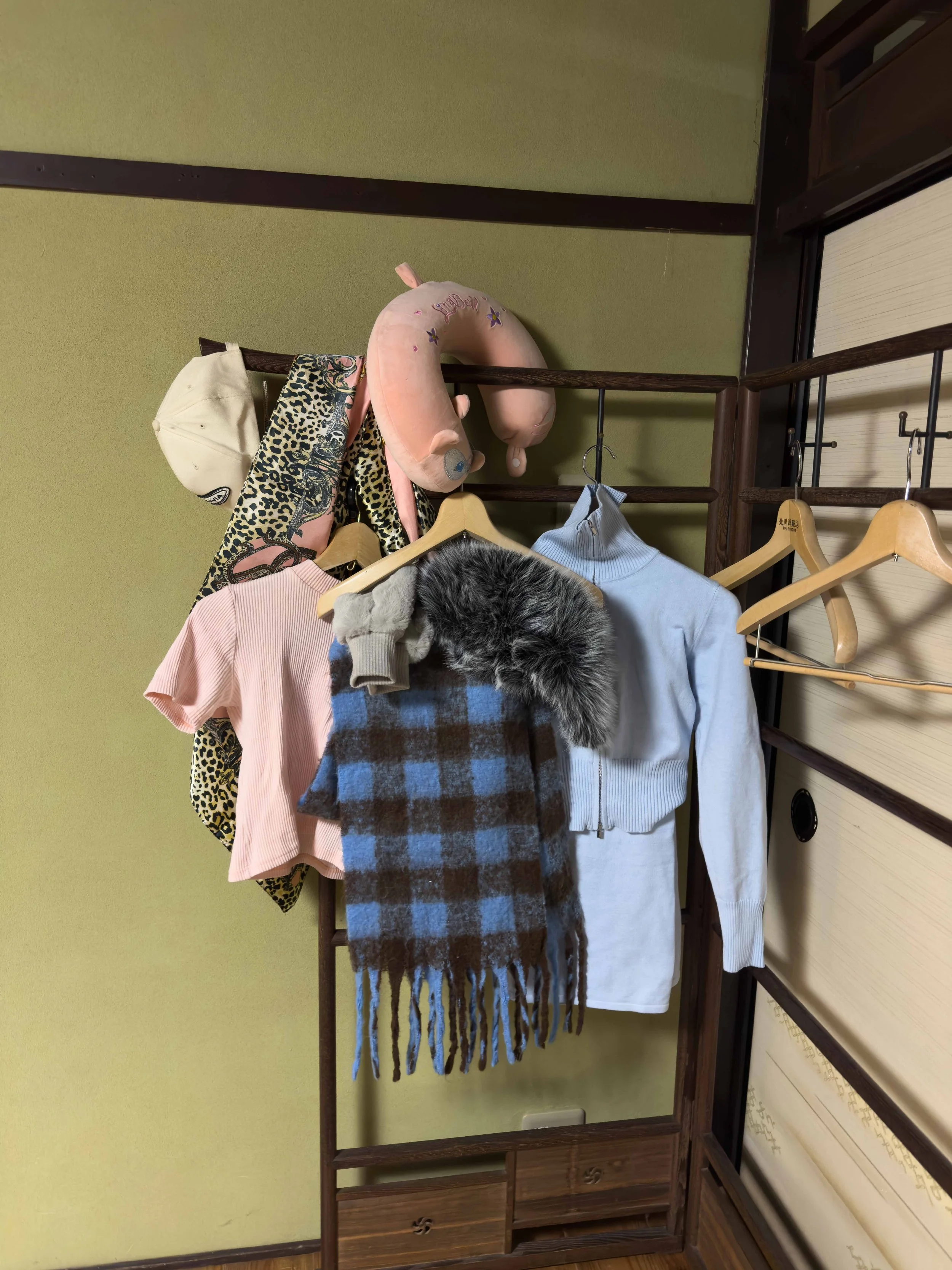 Estantería de madera con ropa colgada, incluyendo suéter blanco, bufanda a cuadros morada y azul, gorra beige, y una bufanda con leopardo y detalles en rosa. En la parte superior hay un peluche de luna con caras y globos decorativos.