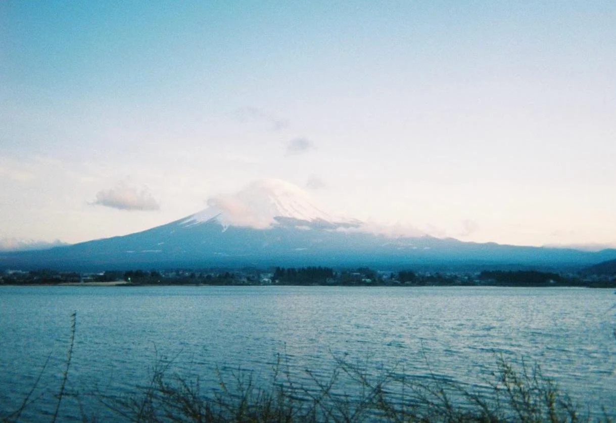 Vista de un volcán nevado, posiblemente el Monte Fuji, visto desde un cuerpo de agua con vegetación en primer plano, durante el amanecer o atardecer.