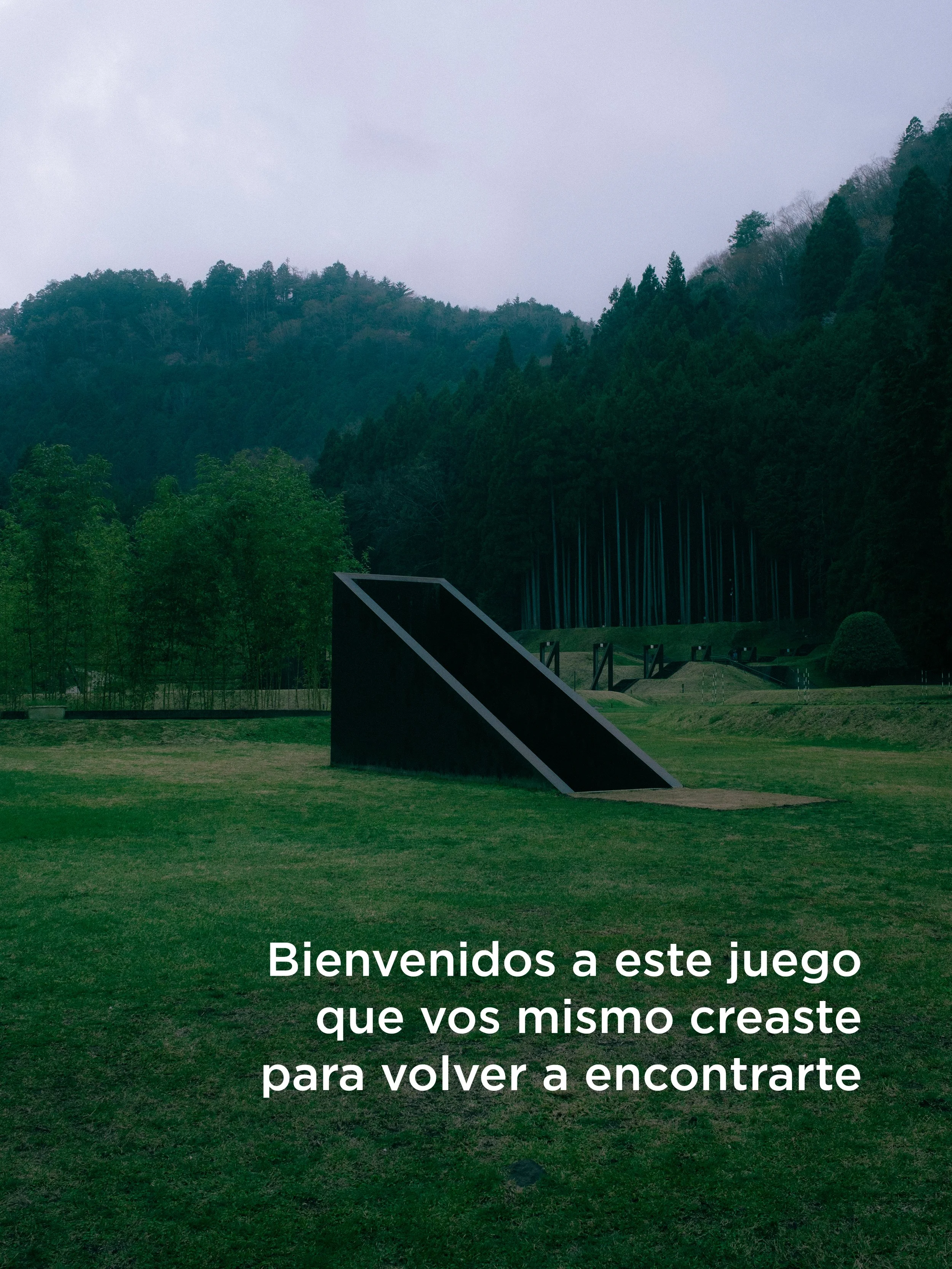 Un campo de césped con colinas y árboles en el fondo, en un día nublado. En la parte inferior de la imagen hay un símbolo en forma de triángulo y escrito que dice: "Bienvenidos a este juego que vos mismo creaste para volver a encontrarte".