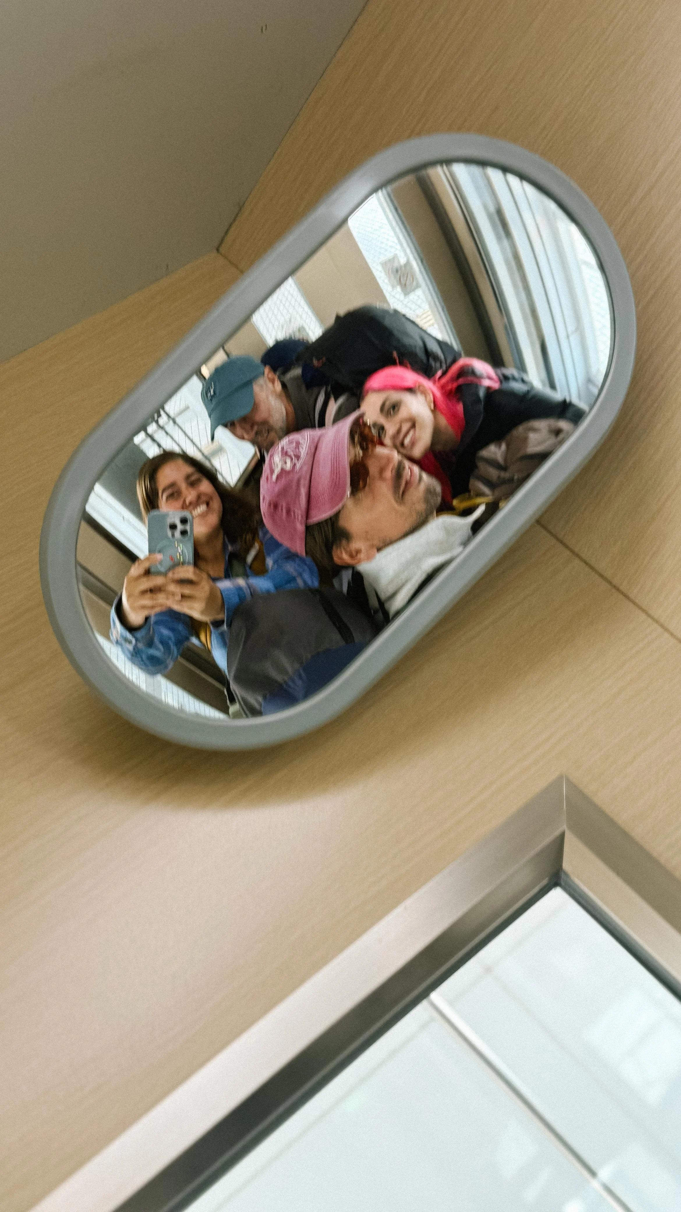 Grupo de personas tomando una selfie en un espejo, con sonrisa, en un lugar con ventanas grandes y estructura metálica.