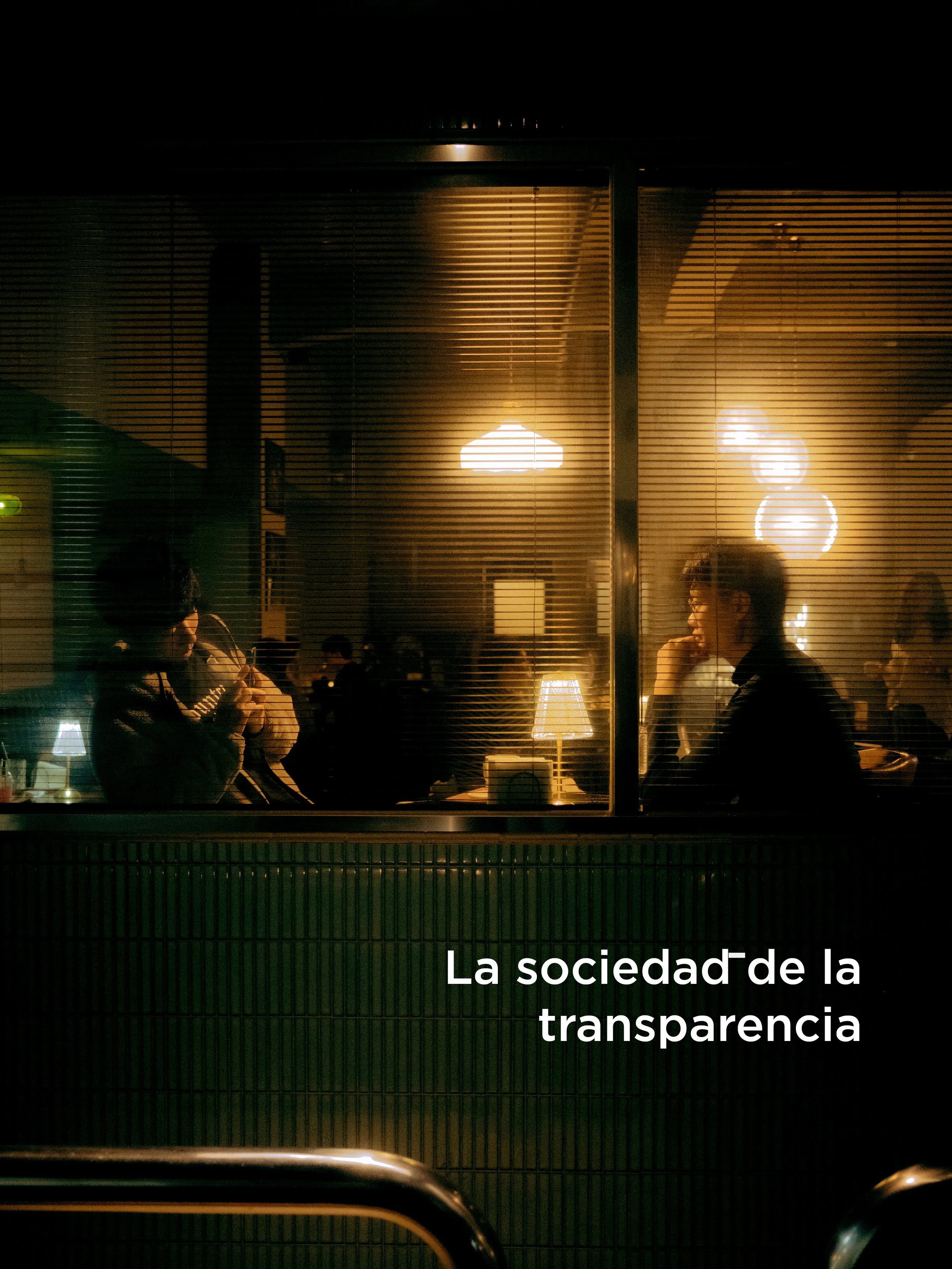 Imagen de una vitrina de un negocio con personas sentadas en el interior, visible a través de las persianas. Hay una lámpara en el interior y las paredes están decoradas con lámparas de luces cálidas.