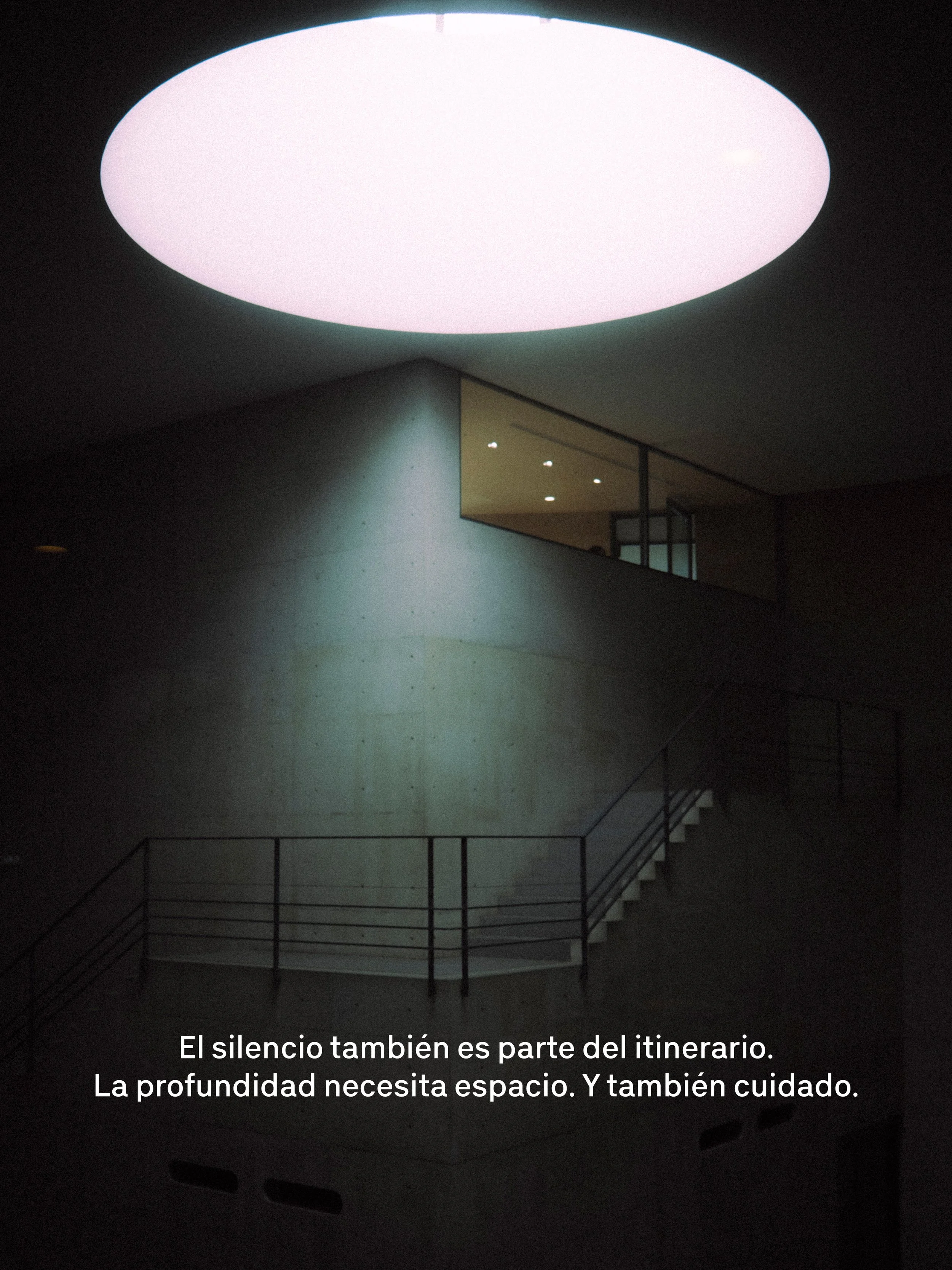 Vista de un espacio interior con un techo luminoso en forma ovalada y una escalera con barandillas de metal. El texto en la imagen dice: "El silencio también es parte del itinerario. La profundidad necesita espacio. Y también cuidado.".