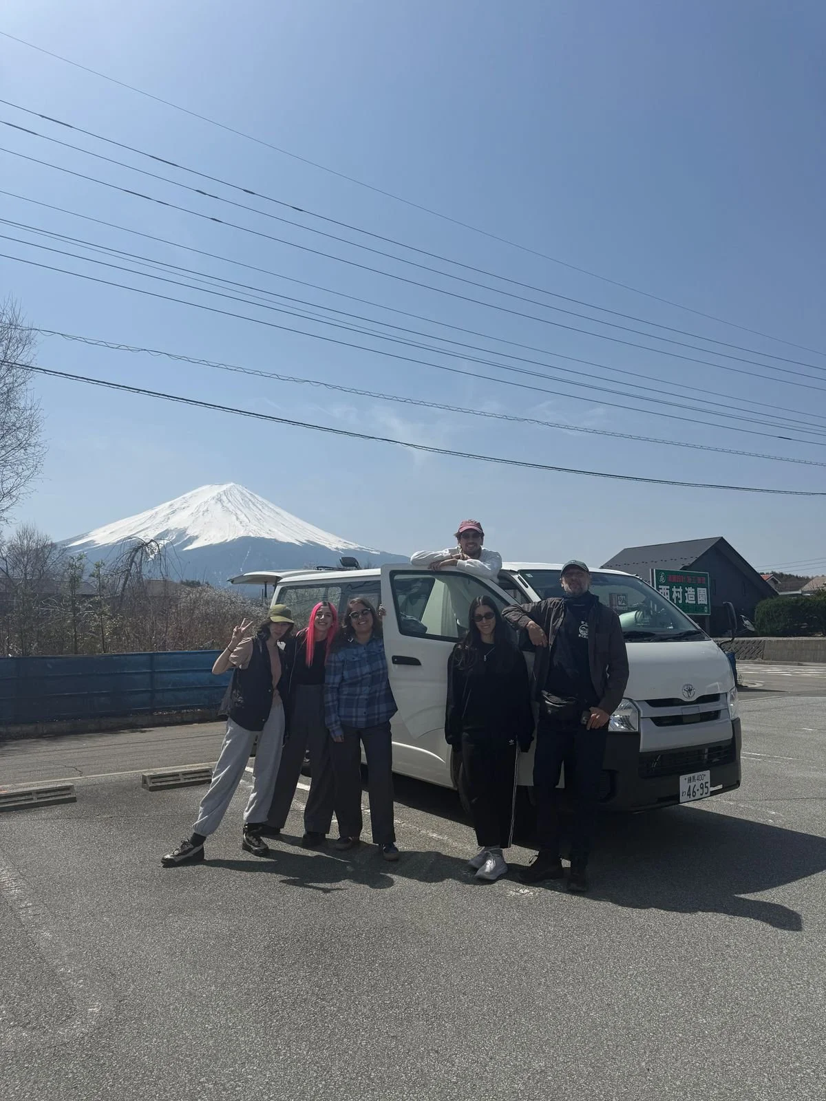 Grupo de personas posando frente a una camioneta blanca con el volcán Fuji en el fondo, en un día soleado.
