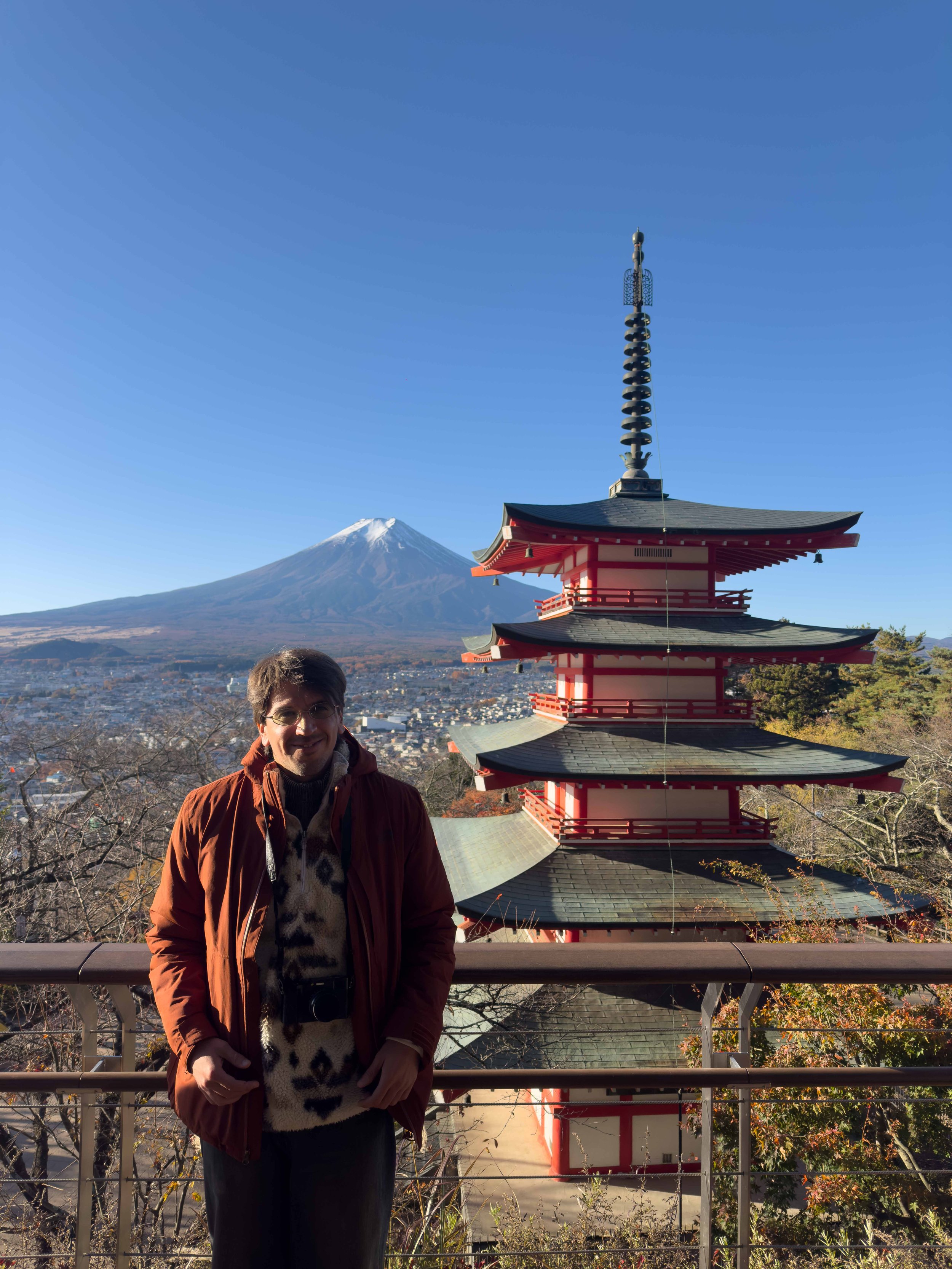 Hombre sonriendo delante de una pagoda japonesa con el volcán Fuji de fondo bajo un cielo despejado.