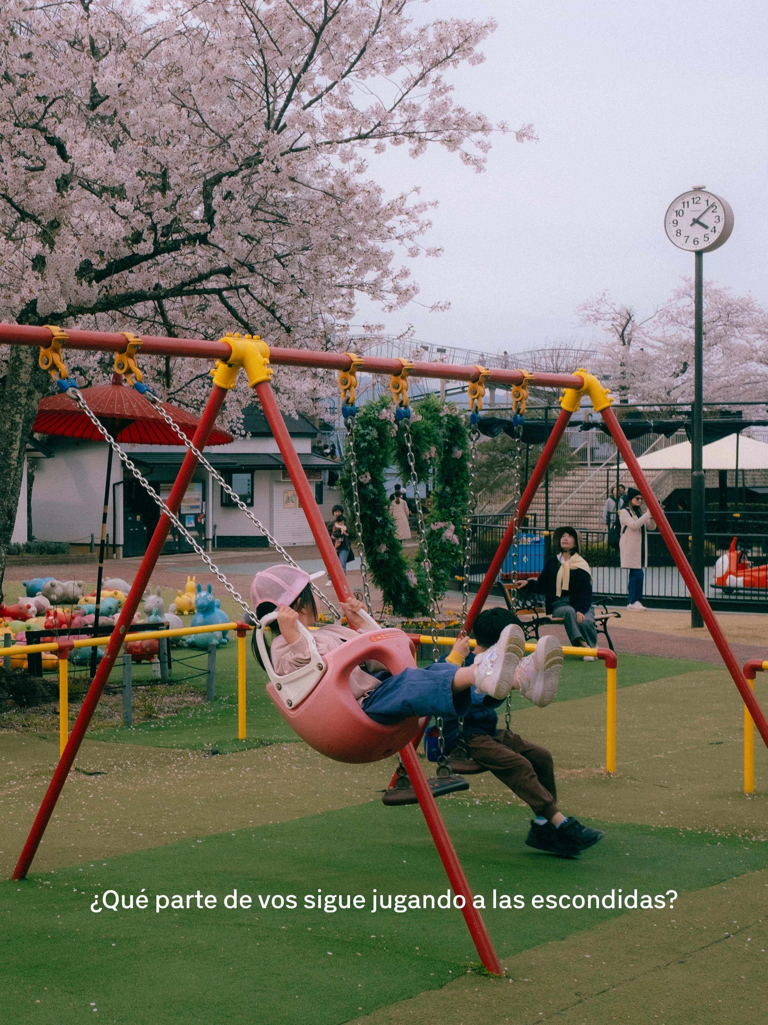 Niños jugando en un parque con columpios en medio de cerezos en flor. Se puede ver la estructura del parque, bancos, una señora sentada en un banco, y varias personas paseando o tomando fotos. El cielo está nublado.