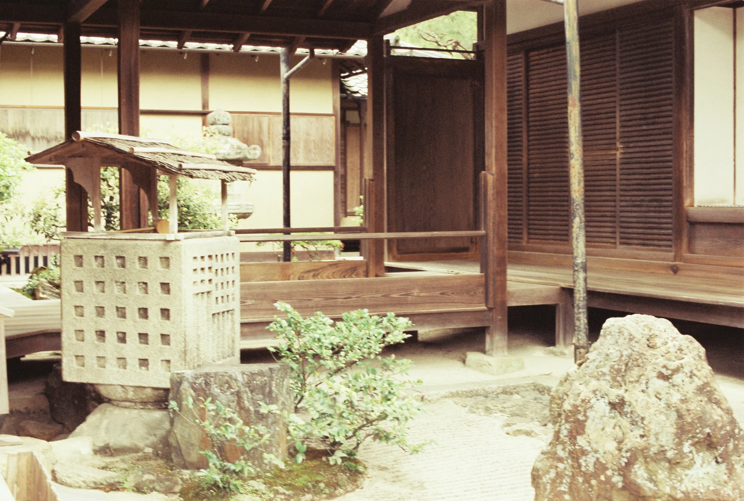 Estructura de madera en un jardín japonés con plantas, piedras y un pequeño pozo de agua.