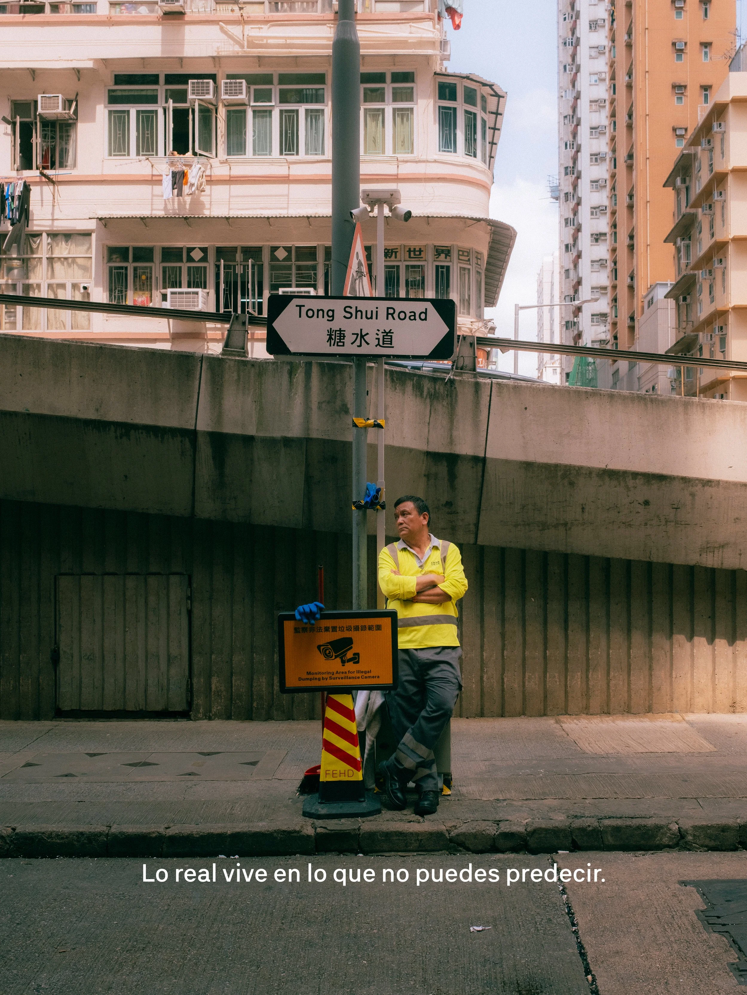 Un hombre de pie en una esquina de la calle, con los brazos cruzados, frente a una señal que indica la calle Tong Shui Road. Hay un cartel que advierte sobre monitoreo para lanzar basura ilegal con cámaras de vigilancia. En el fondo, edificios residenciales de múltiples pisos y un cartel en chino. La frase en la parte inferior de la imagen dice: "Lo real vive en lo que no puedes predecir."