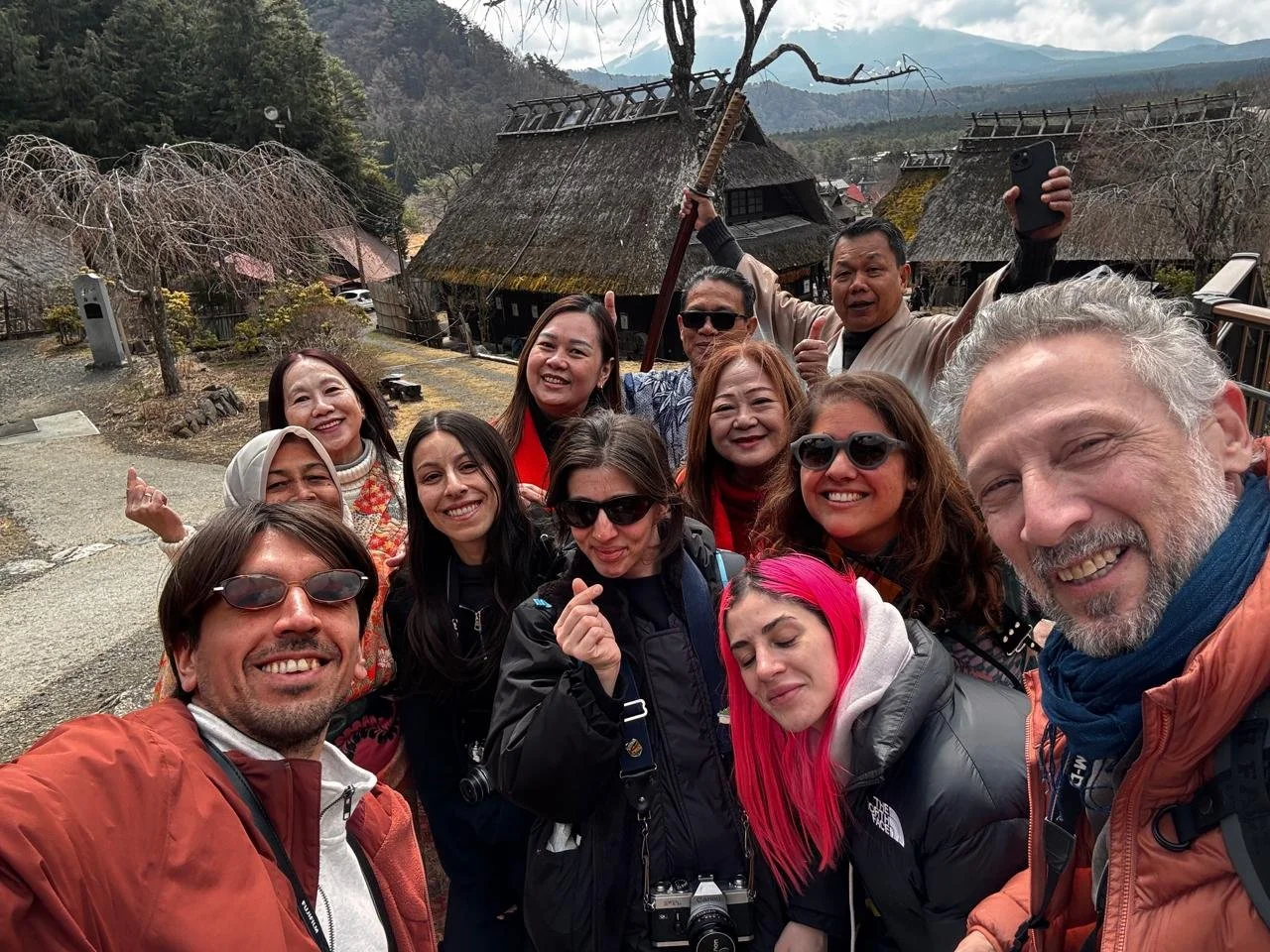 Grupo de personas sonriendo para una selfie en un entorno rural con casas tradicionales de techos de paja y árboles sin hojas, en un paisaje montañoso.
