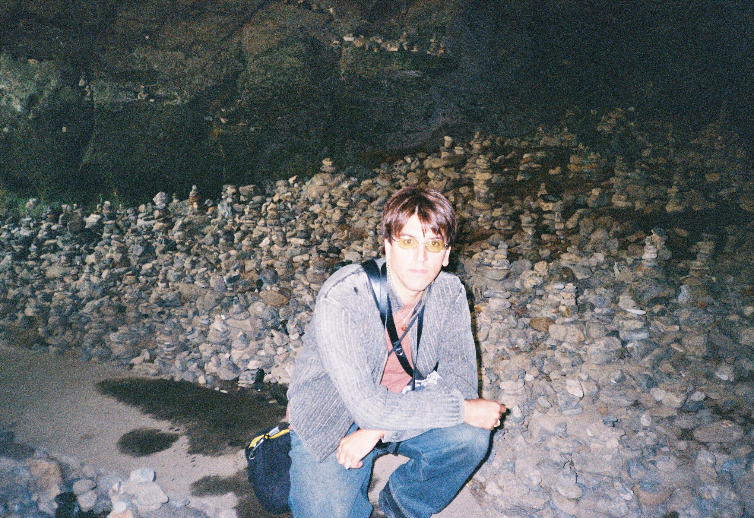 Persona con gafas de sol, camiseta de manga larga gris, pantalón azul, mochila, en un entorno de cueva con muchas rocas y estalactitas.