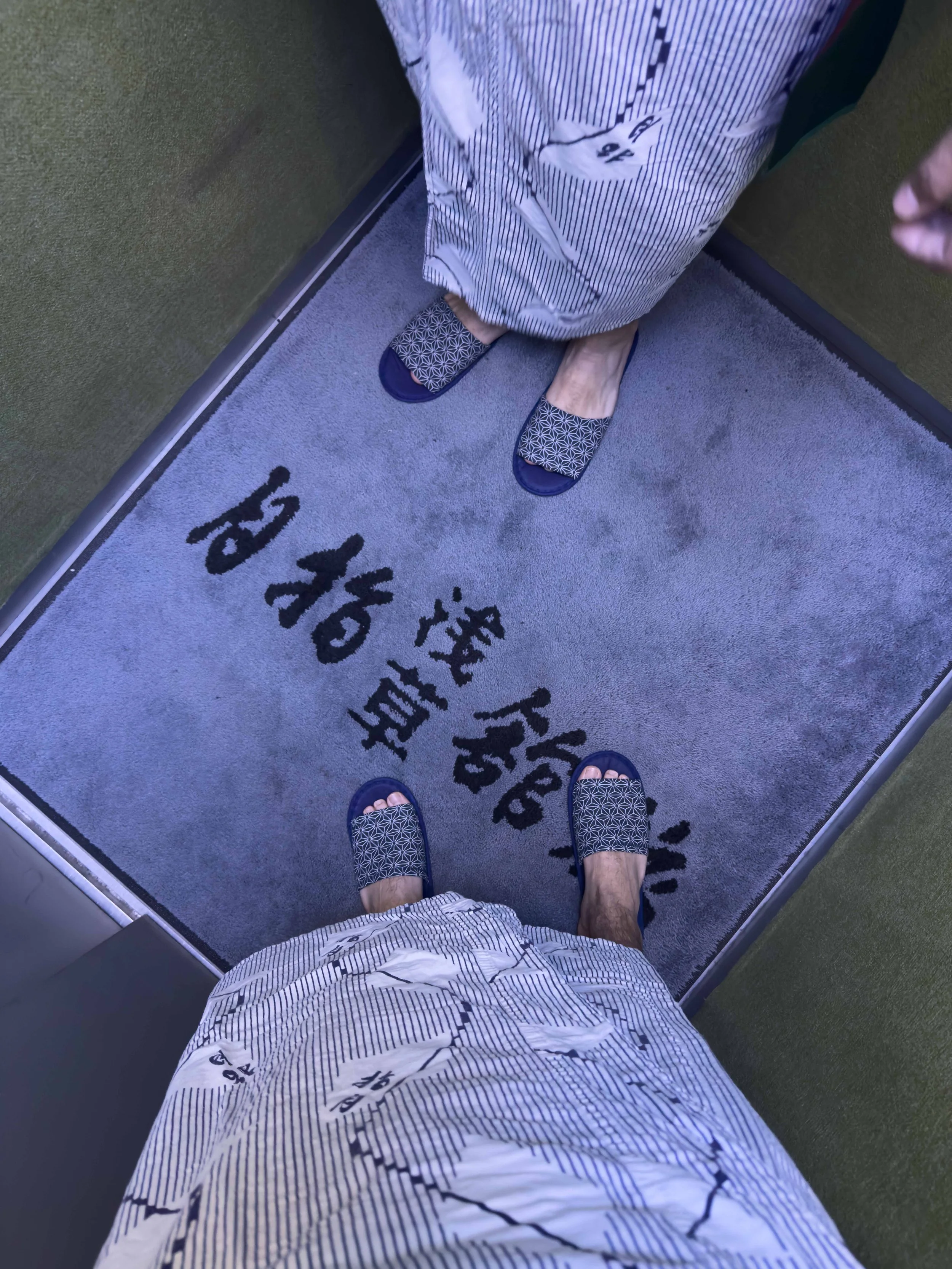 Vista del suelo de una cabina de ascensor con una alfombra gris y caracteres en japonés, desde la perspectiva de una persona que lleva sandalias con patrón geométrico y un piyama de rayas blancas con negro.