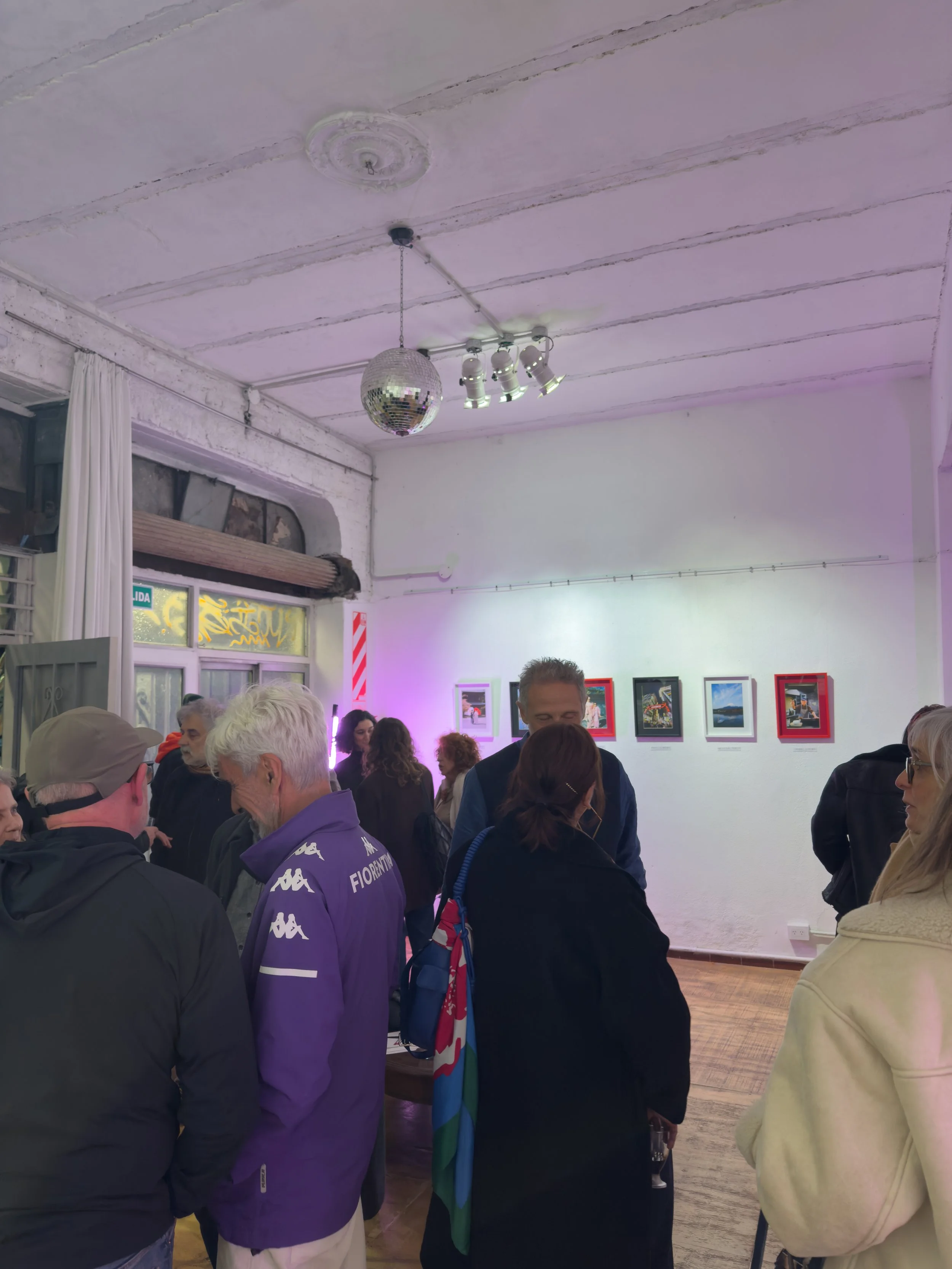 Una muestra de personas en una galería de arte, observando fotografías enmarcadas en una pared blanca, con un ambiente de iluminación suave y decoraciones en el espacio.