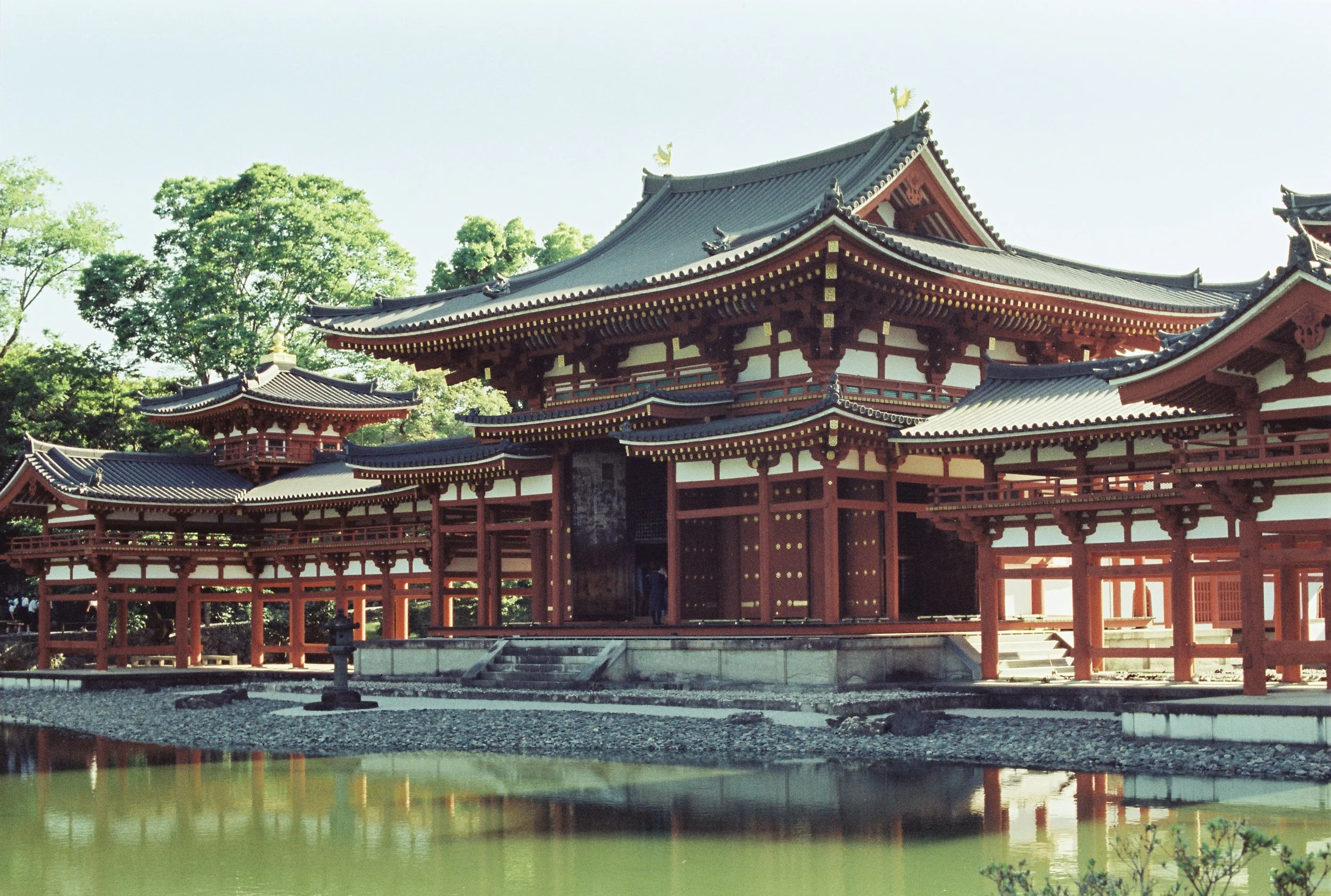 Un templo tradicional japonés con techos de tejas y estructura de madera pintada de rojo, rodeado de árboles verdes y reflejado en un estanque en primer plano.
