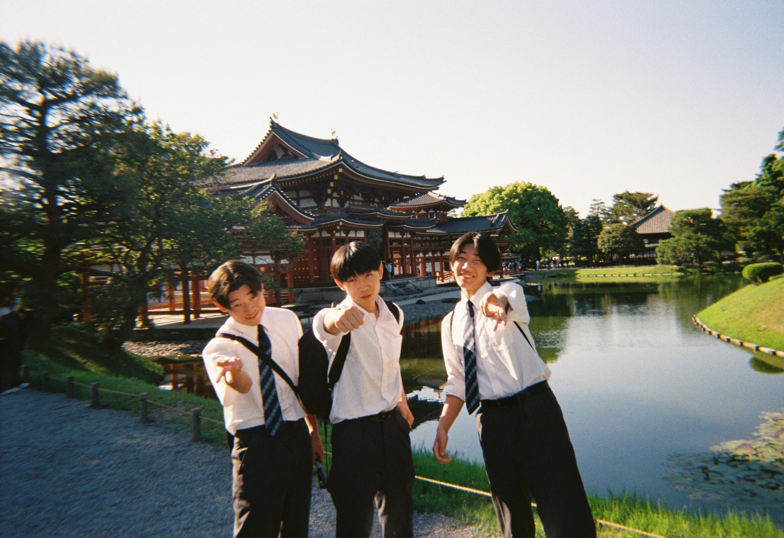Grupo de adolescentes con uniforme escolar posando junto a un estanque en un parque con arquitectura tradicional japonesa y árboles verdes en el fondo.