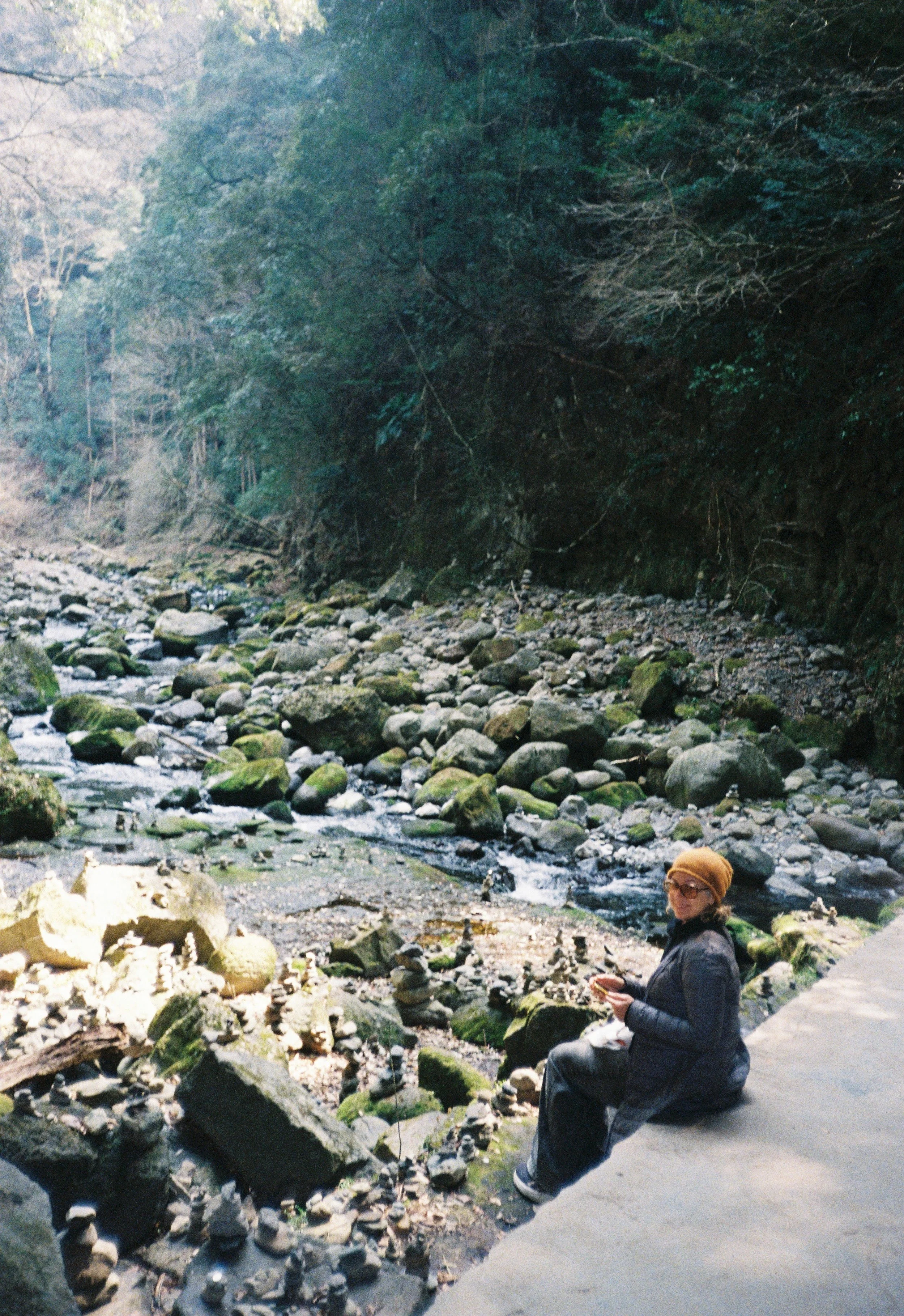 Persona sentada junto a un río rodeado de rocas, en un entorno boscoso, con árboles altos y vegetación, vistiendo gafas, abrigo y gorro, sosteniendo una taza.