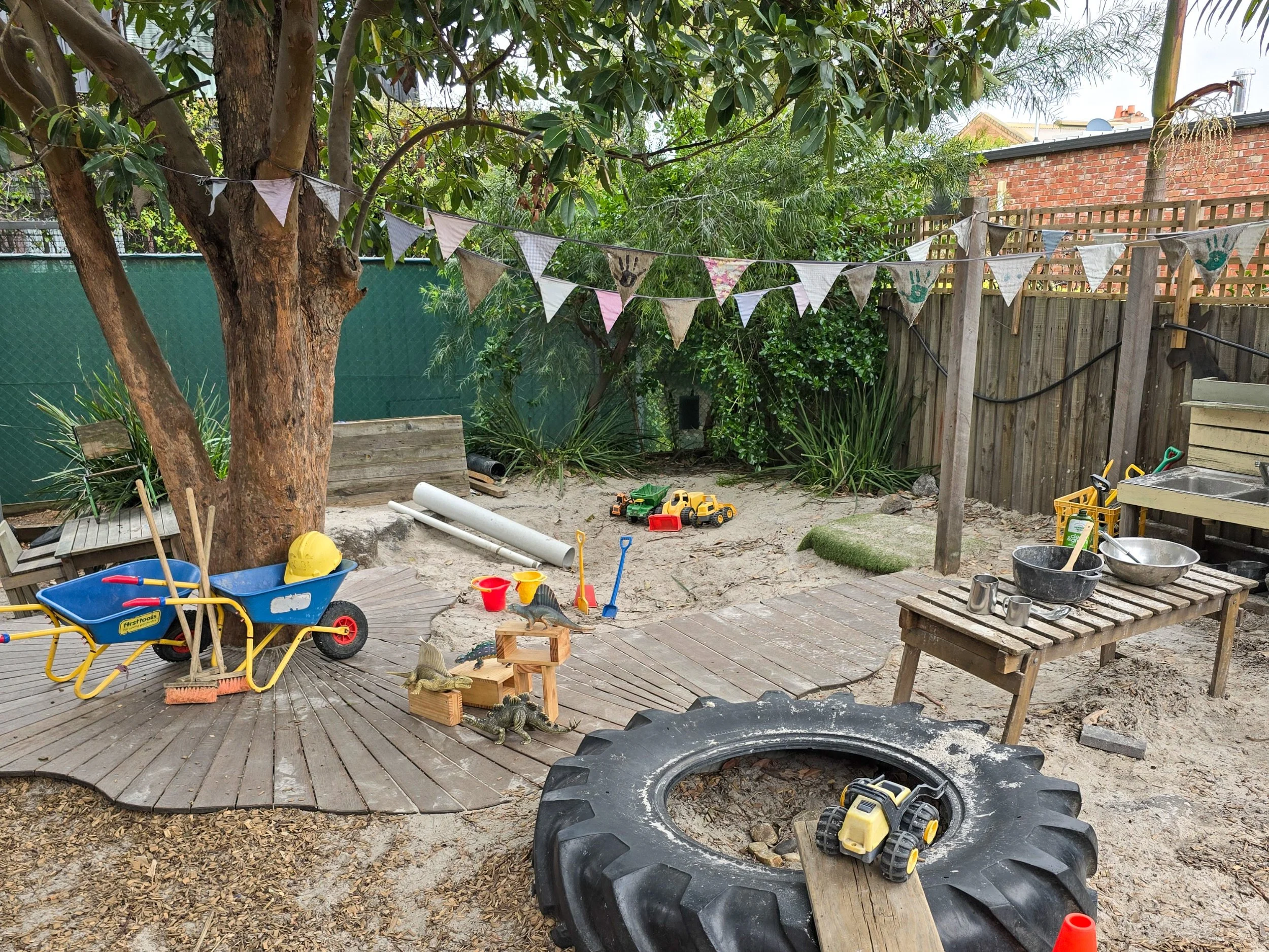 Outoor play area with a large tree, sandpit, tyre and lots of toys