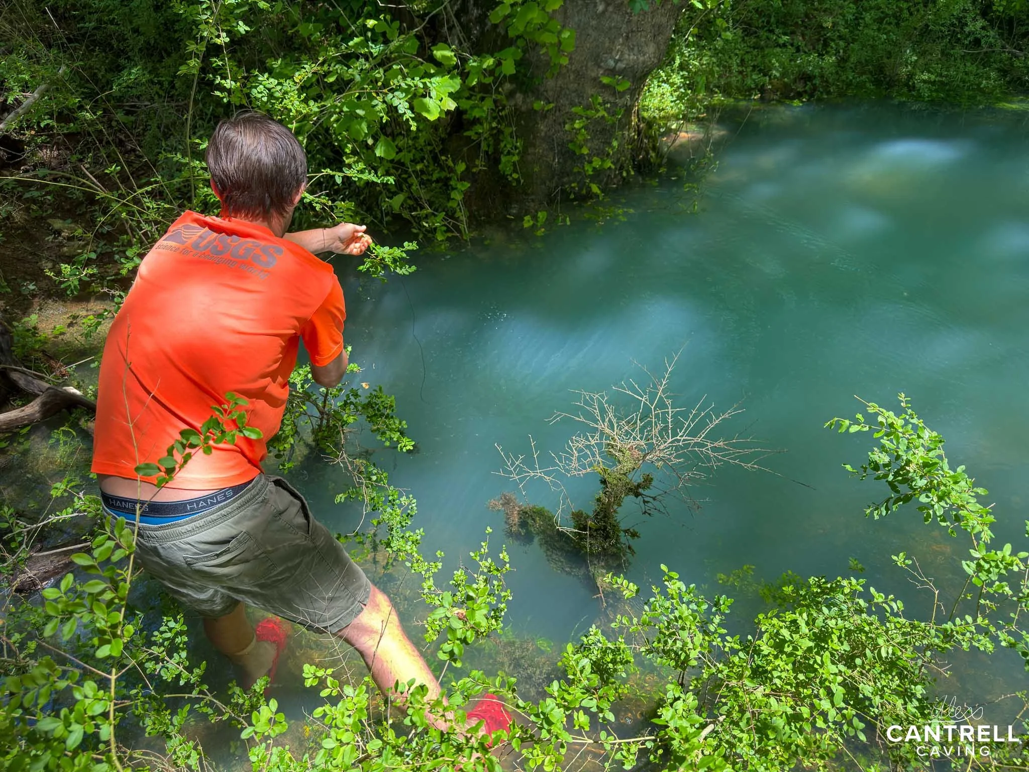 A person wearing an orange shirt standing near a blue-green body of water, surrounded by lush green vegetation, in a nature setting.