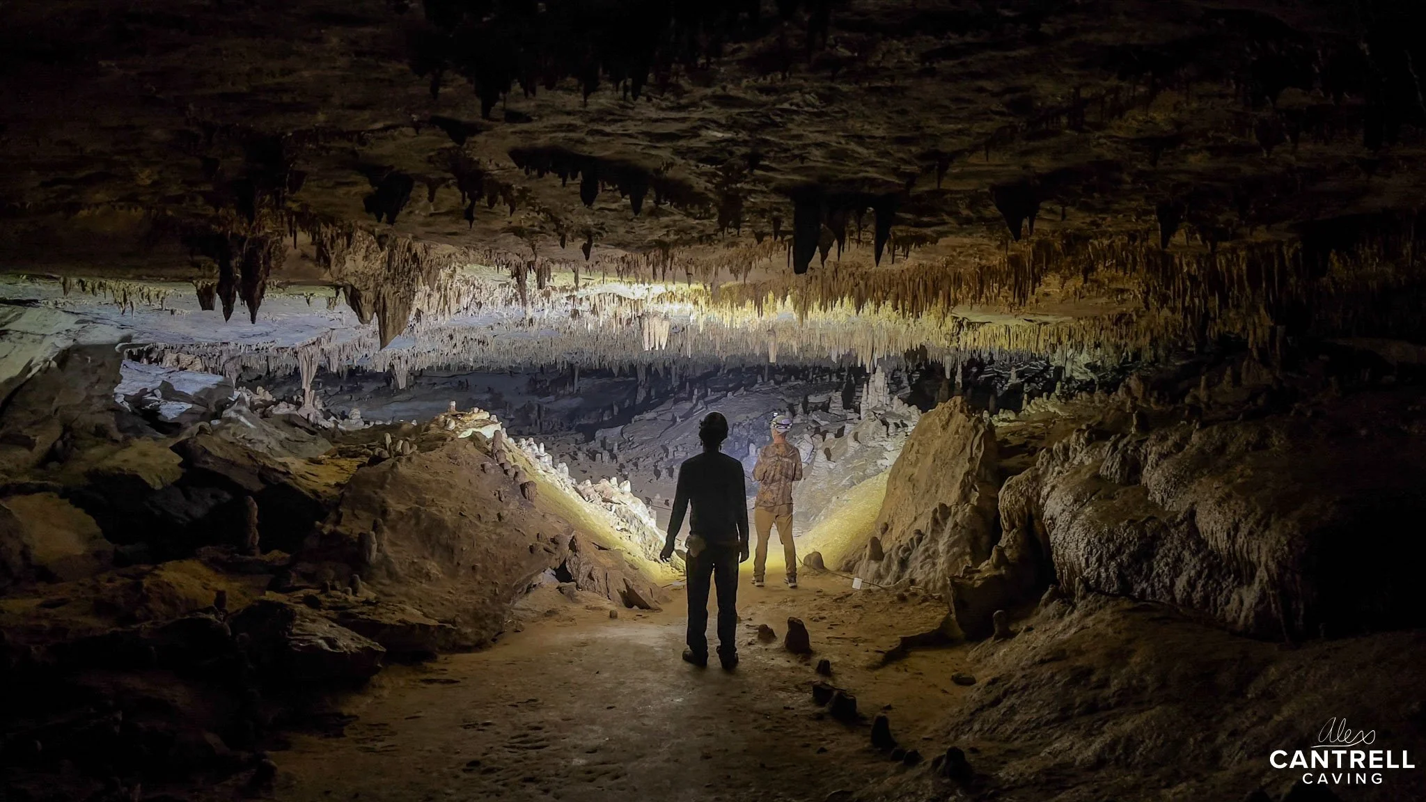 Two people exploring a cave with stalactites and stalagmites illuminated by flashlight.