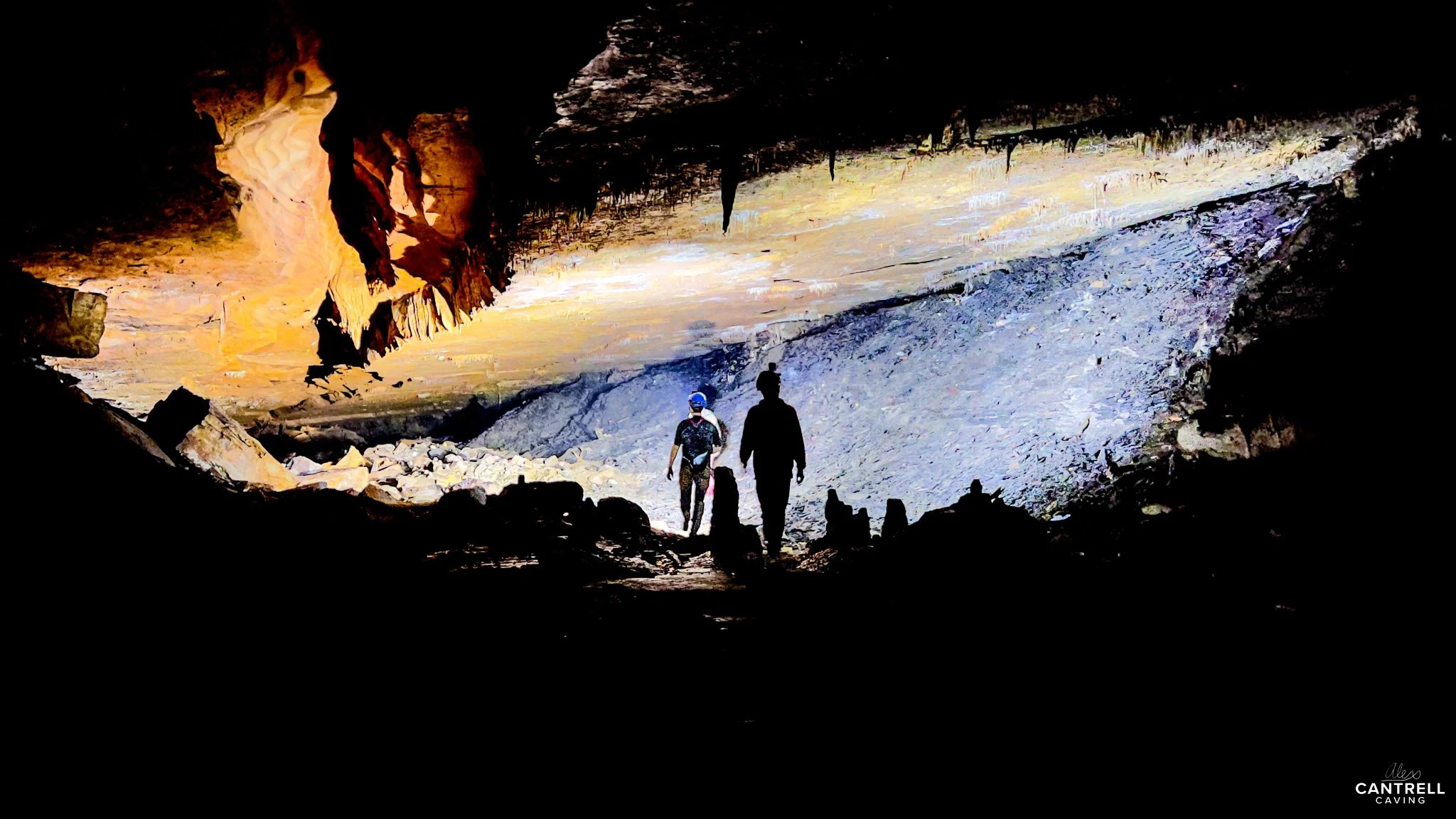 Cave interior with two people wearing helmets standing on rocky ground. Illuminated rock formations and stalactites visible on the ceiling.