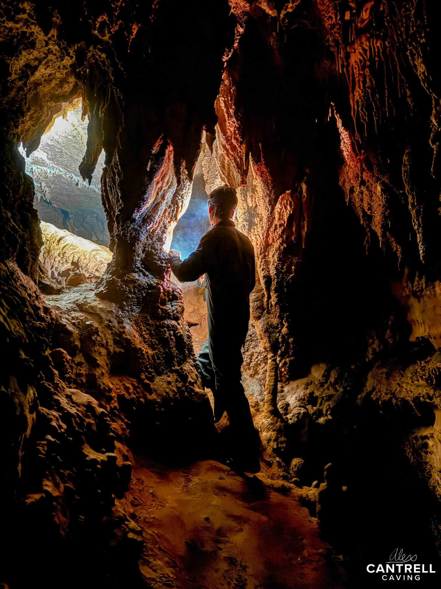 A person explores a cave with stalactites and rock formations, lit by natural light from an opening.