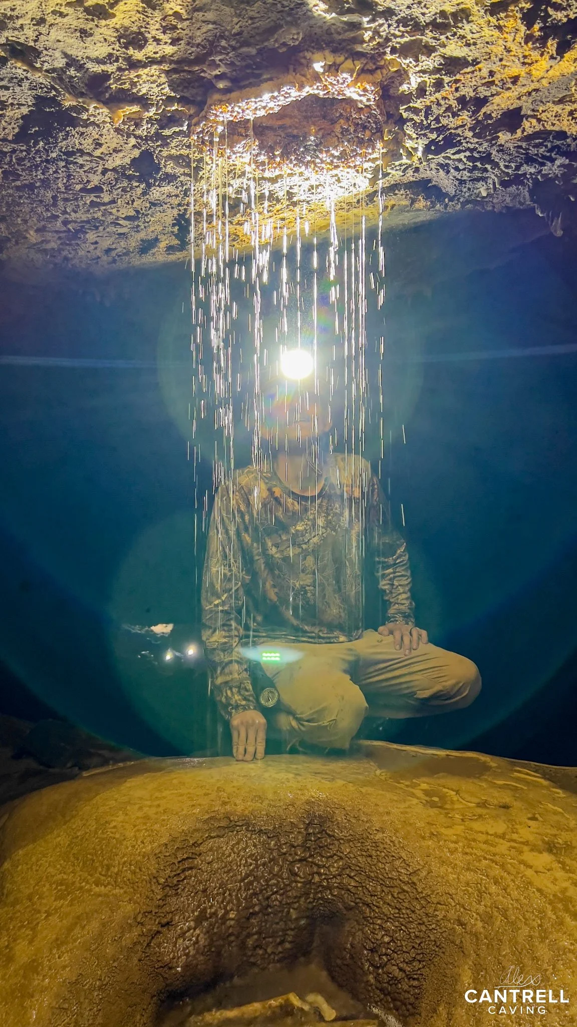 Person wearing a headlamp in a cave, with water dripping from the rocky ceiling.