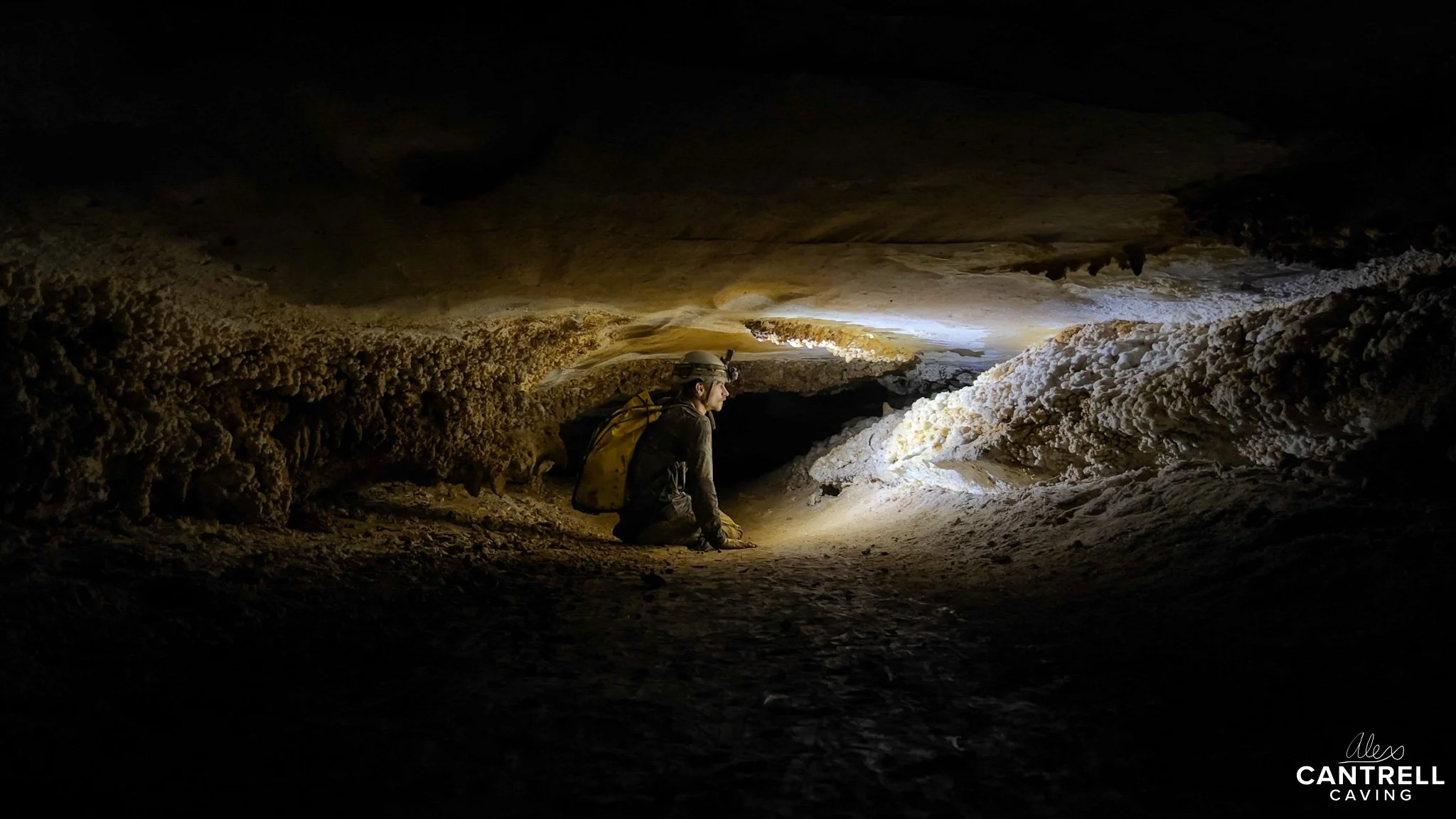 Person with helmet exploring a narrow cave passage, equipped with a headlamp, carrying a backpack. The cave walls are rough and textured, with shadows highlighting the formation features.