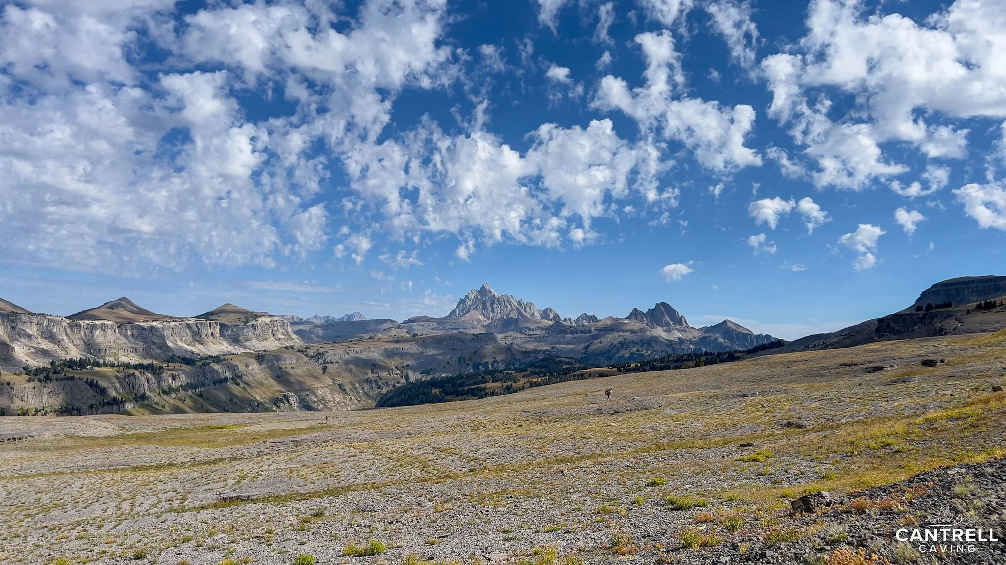 Barren landscape with distant rocky mountains under a blue sky with clouds, featuring scattered vegetation and humans in the foreground.