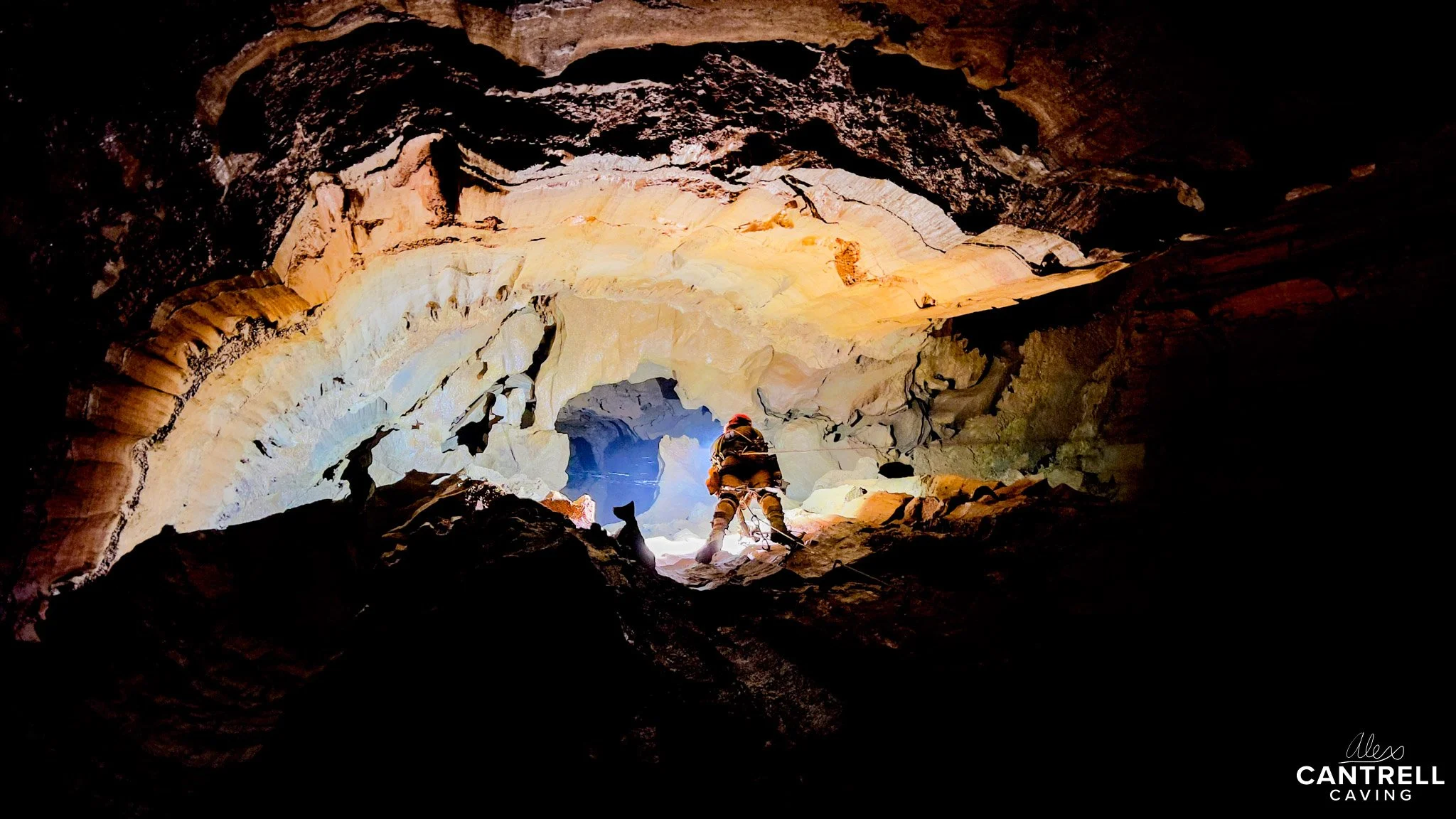 Person exploring a brightly lit cave, surrounded by rock formations, seen from behind. "Cantrell Caving" logo in the corner.