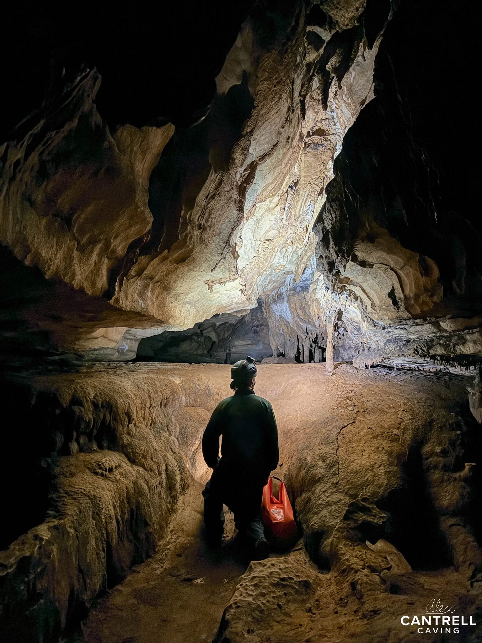 A person in caving gear kneels inside a dimly lit cave, with a backpack beside them. They are facing a large, illuminated rock formation.