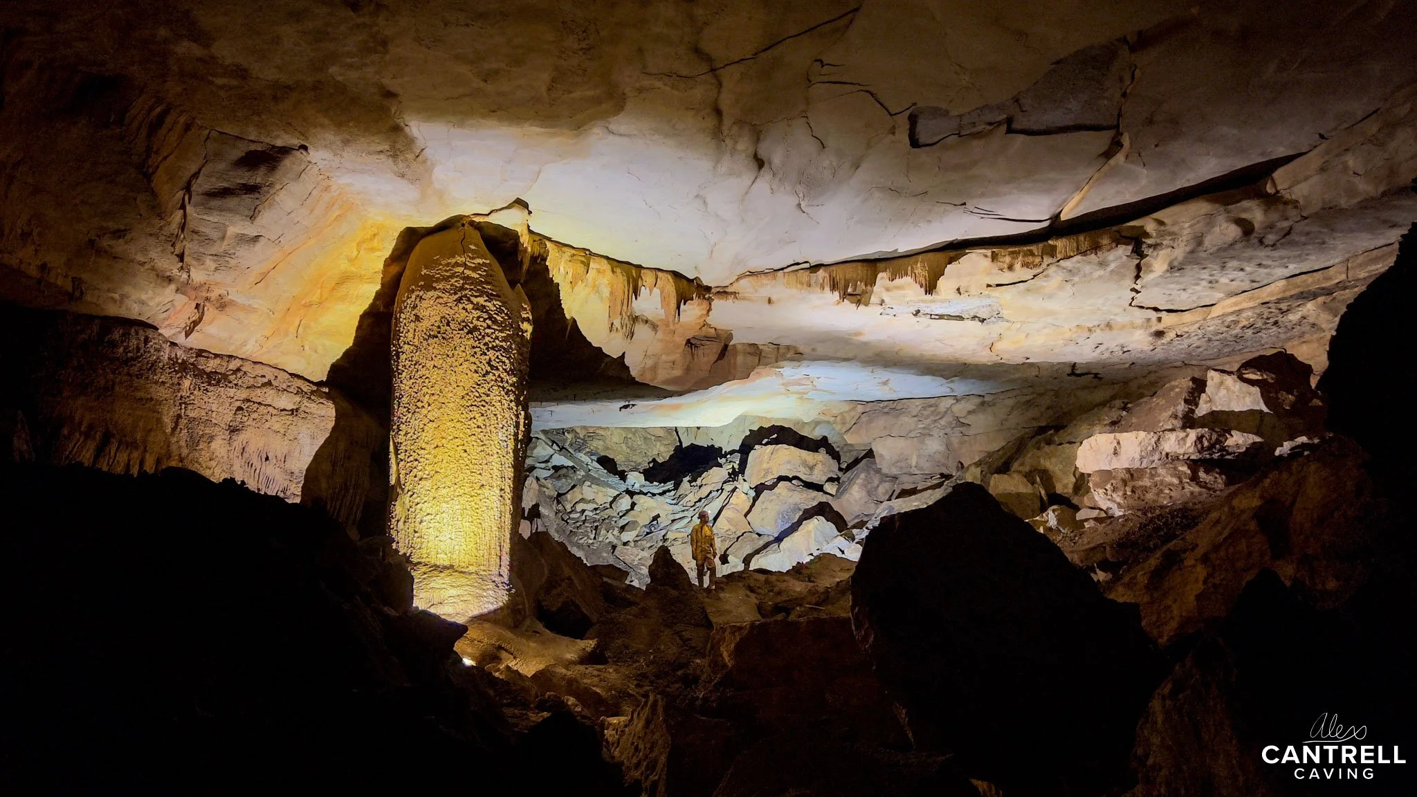 Interior of a cave with stalactites and stalagmites, rocky formations, and a person exploring. The cave is illuminated by artificial light.