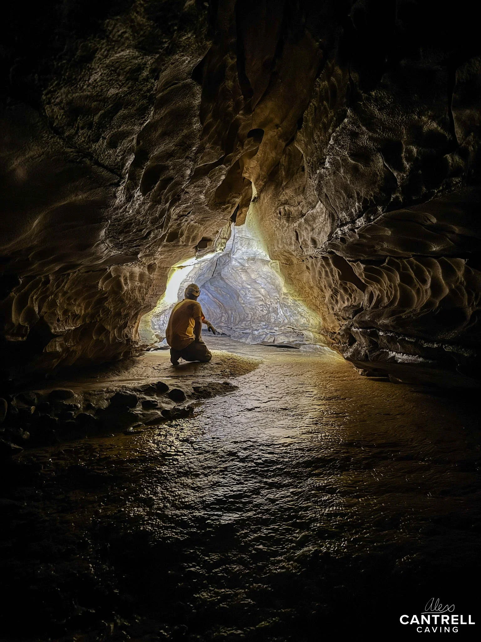 Person exploring inside a dimly lit cave, kneeling on a rocky surface. Light shines in from an opening, illuminating textured cave walls.