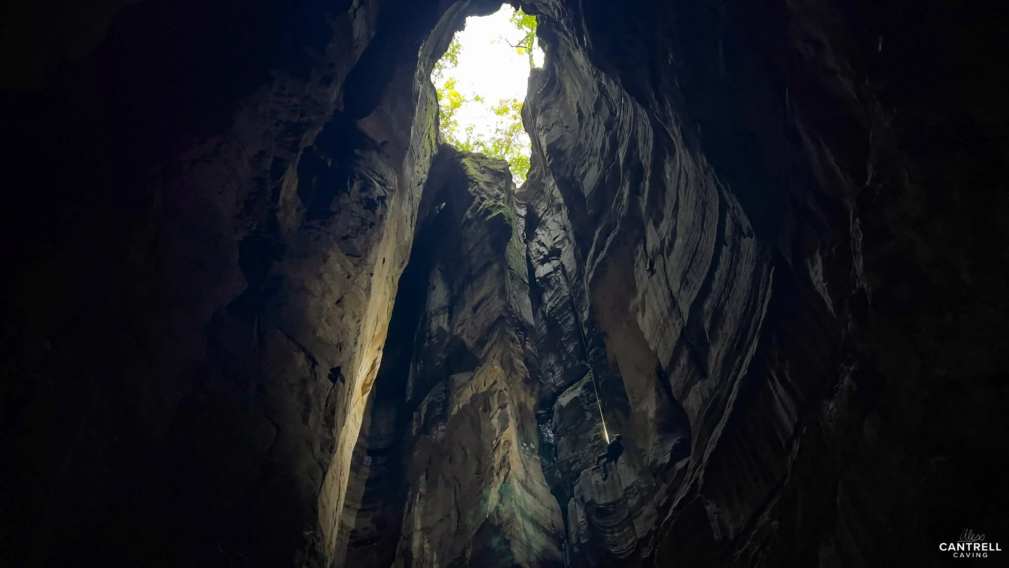 Interior view of a cave with light entering from an opening at the top, illuminating natural rock formations. A person is rappelling down one side of the cave wall.