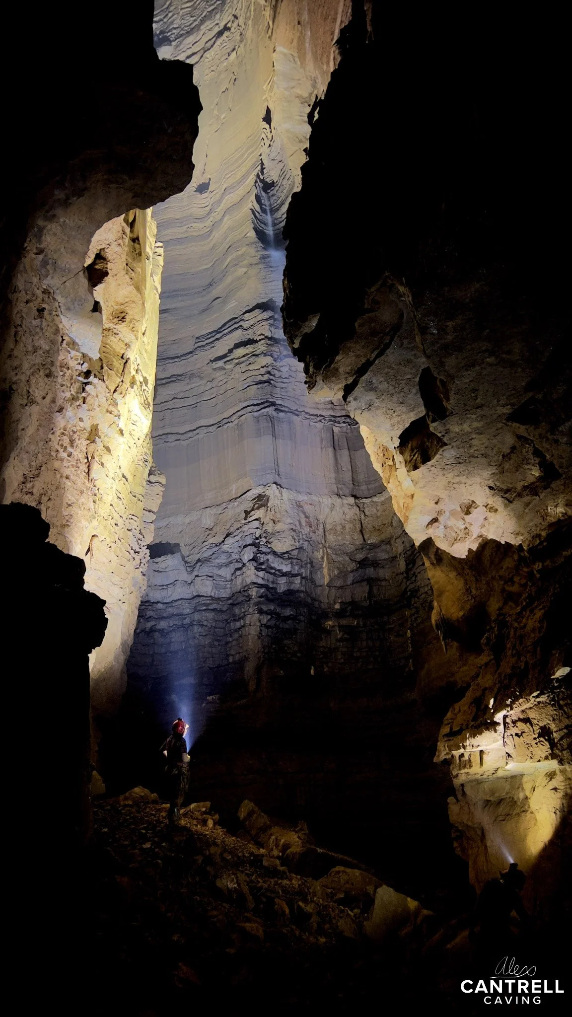 Person exploring a large, illuminated cave with rugged rock formations and tall ceilings, holding a flashlight.