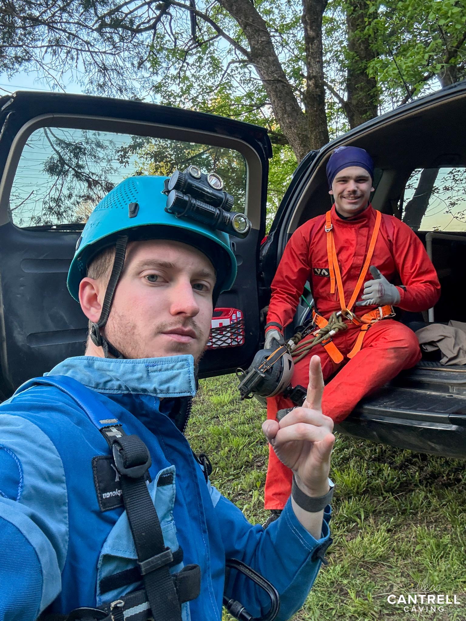 Two people in caving gear outside a vehicle, with trees in the background. One person is wearing a blue helmet with a headlamp, while the other sits in the back of a vehicle wearing red overalls.