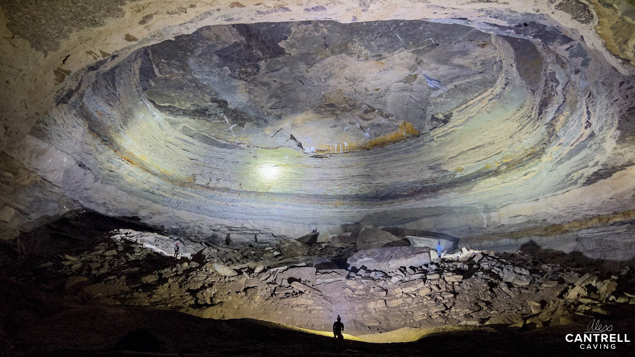 Large underground cave with layered rock formations and scattered boulders, illuminated by lights. A person is visible in the foreground for scale.