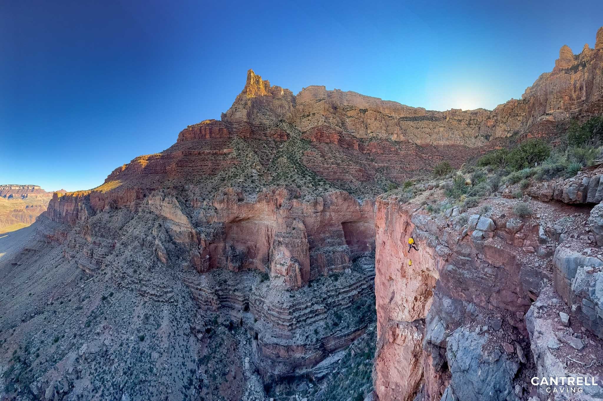 A rock climber scaling a red rock cliff in the Grand Canyon under a clear blue sky. Rugged terrain and layered rock formations in the background.