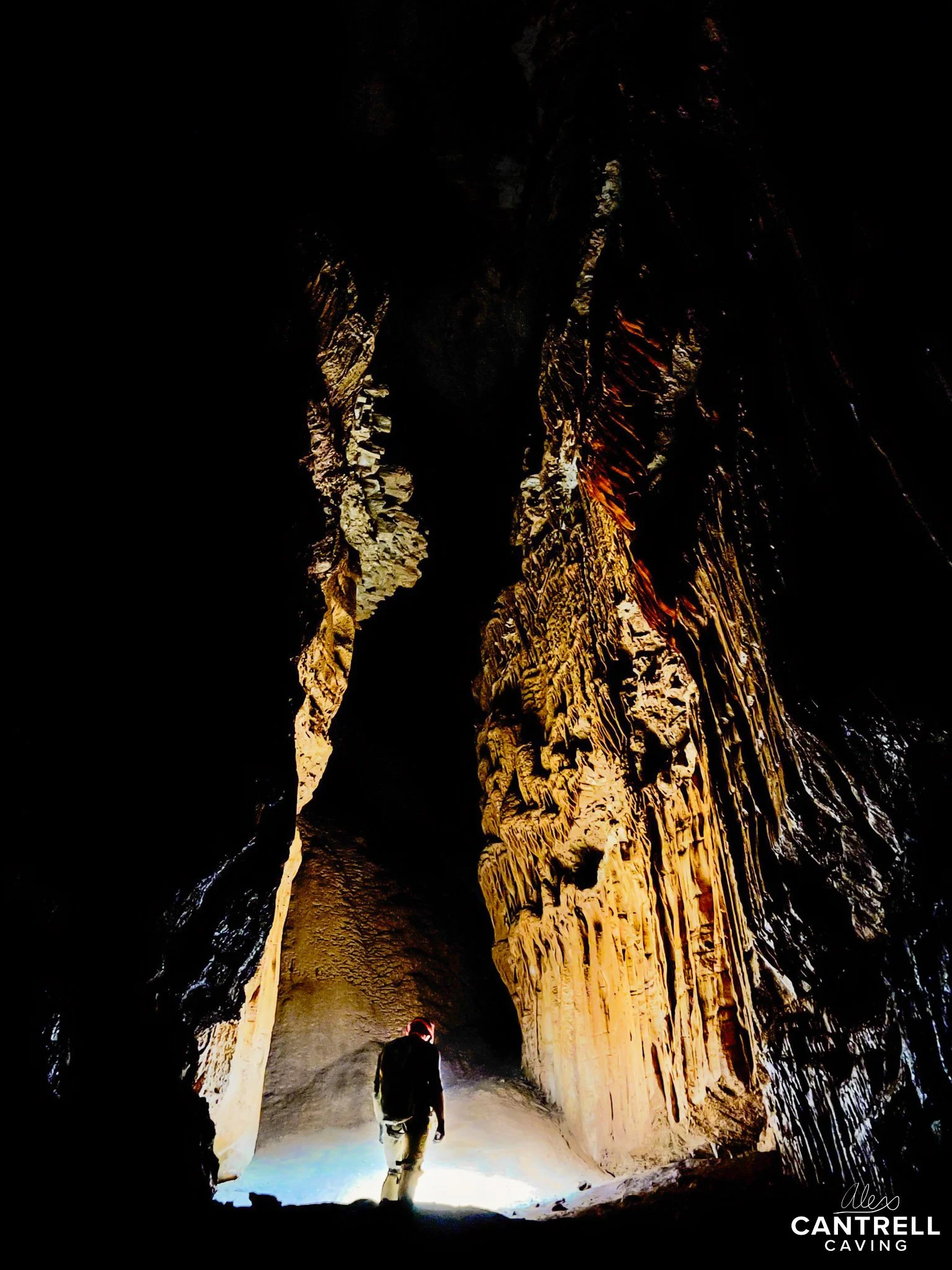 A person stands inside a dimly lit cave, surrounded by towering rock formations. The light creates dramatic shadows on the cave walls, highlighting the textures. The person is facing a bright light source, which casts a glow on the cave floor. There'