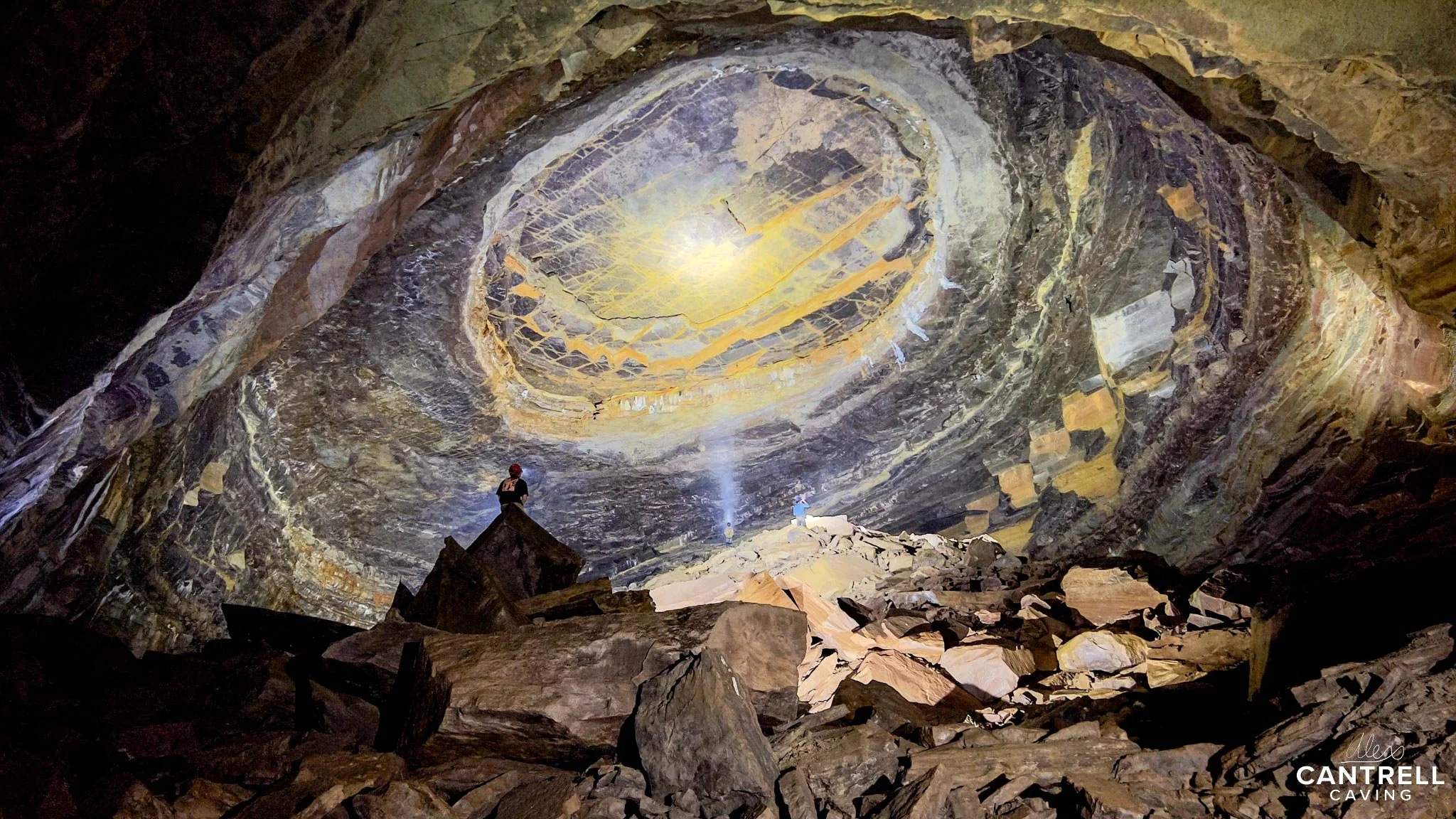 Cavers exploring a large underground cave with a rocky ceiling feature resembling a dome and scattered stone debris on the ground.