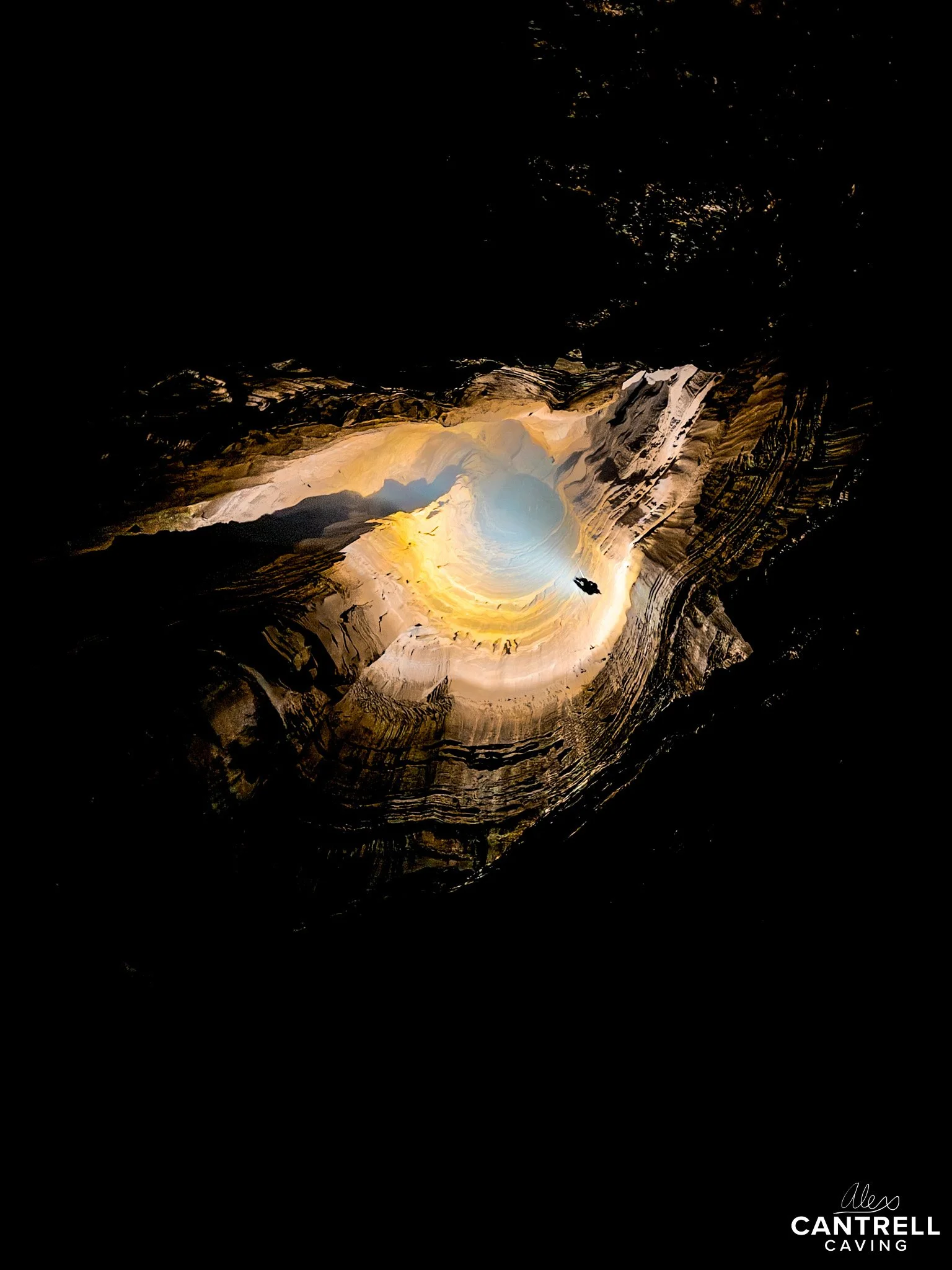 Aerial view of a brightly lit cave or sinkhole with layered rock formations. The light creates a contrast, highlighting the circular shape and geological features.
