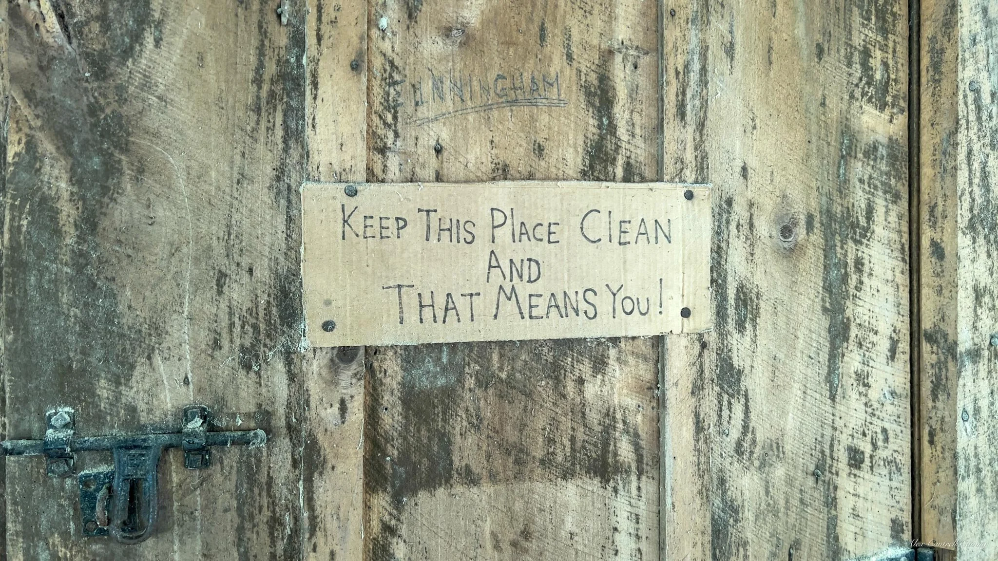 A rustic wooden door with a handwritten sign that reads "Keep This Place Clean And That Means You!" attached to it. The door has a metal latch, and the wood shows signs of wear and age.
